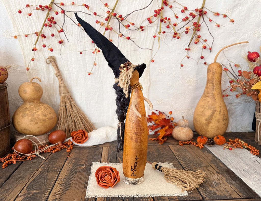 A decorative carved gourd Halloween witch decoration displayed on a rustic wooden table, surrounded by autumn props including a gourd, a straw broom, orange berries, and fall leaves against a white textured backdrop.