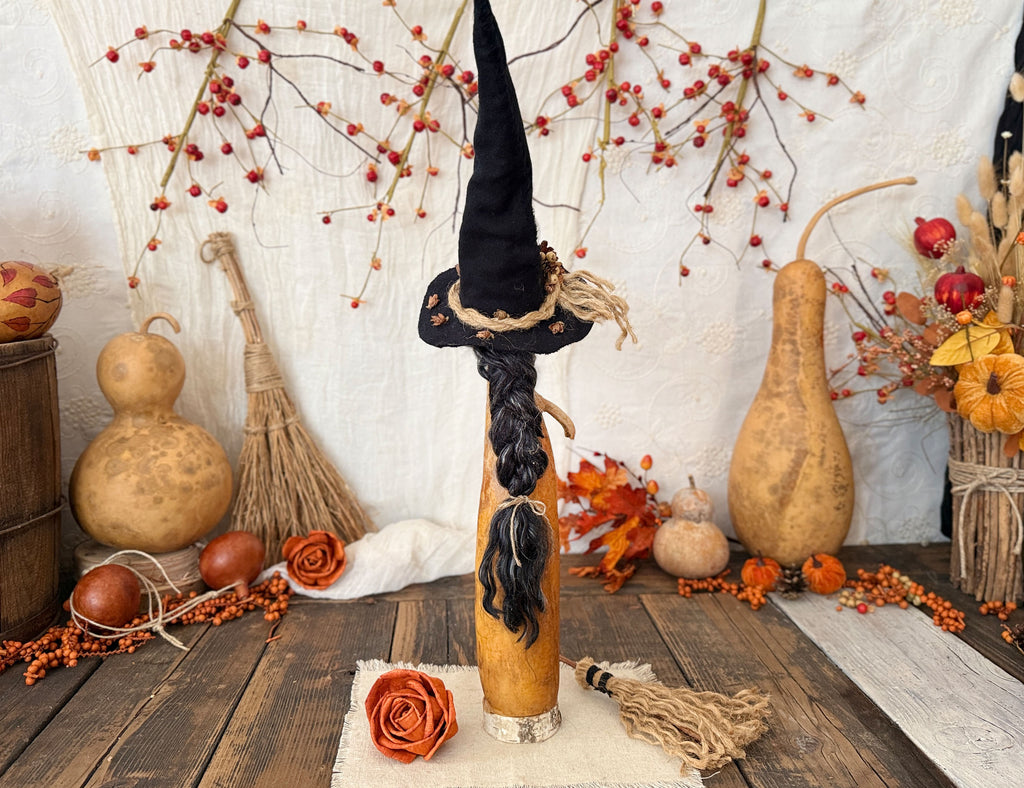 A decorative autumn-themed witch figure with a tall black pointed hat, braided black hair, and a beige body, displayed on a rustic wooden table surrounded by gourd decorations, berries, and fall foliage.