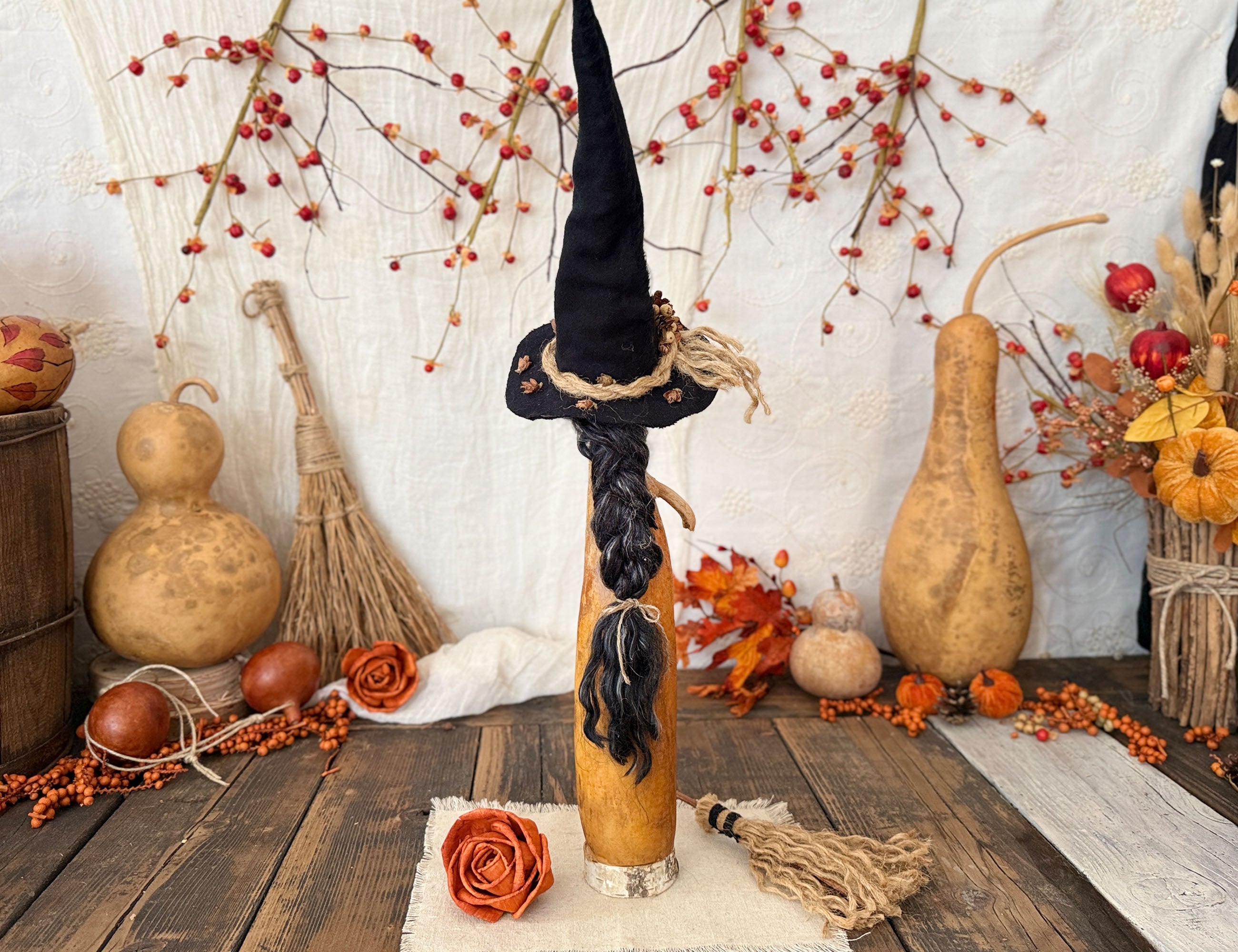 A decorative autumn-themed witch figure with a tall black pointed hat, braided black hair, and a beige body, displayed on a rustic wooden table surrounded by gourd decorations, berries, and fall foliage.