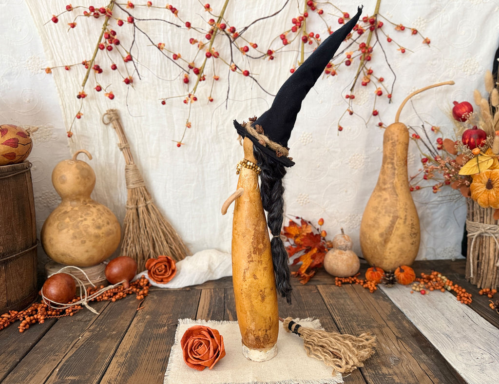 A decorative Halloween display featuring a tall natural gourd figure  with a black witch hat and braided black hair, set on a rustic wooden table with gourd props, berries, and autumn decor.