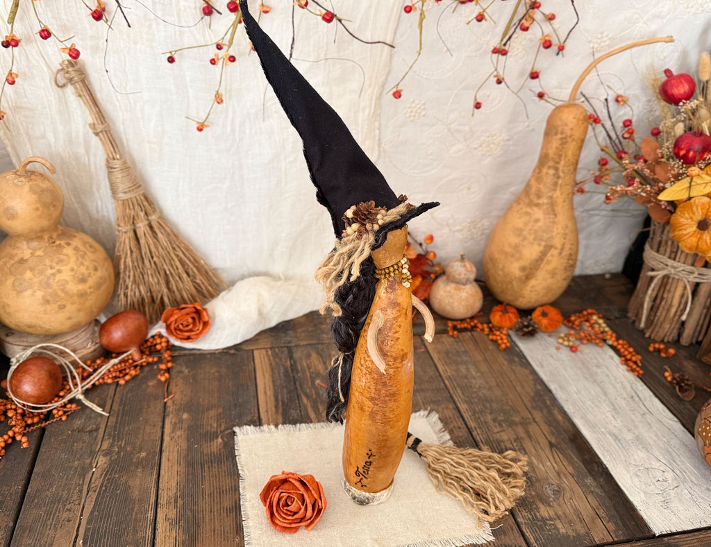 A witch figurine styled with a tall black pointed hat, black yarn hair, and a tassel, set on a rustic wooden surface surrounded by autumn decorations including gourds, dried leaves, and a broom.
