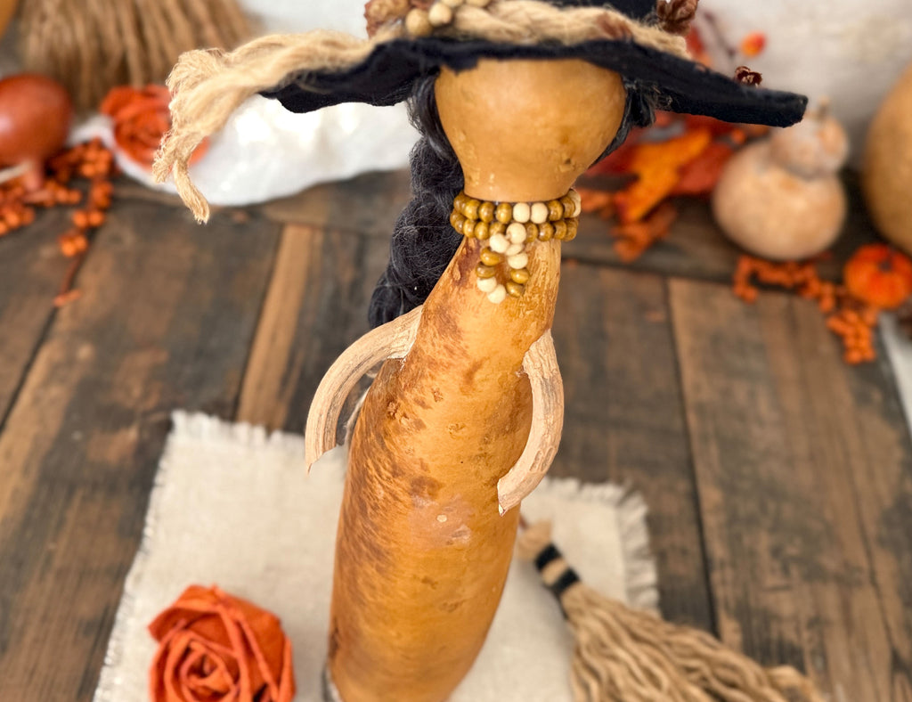 A handcrafted gourd doll with a black hat, braided black hair, and a beaded neck, displayed on a rustic wooden surface with autumn decorations.