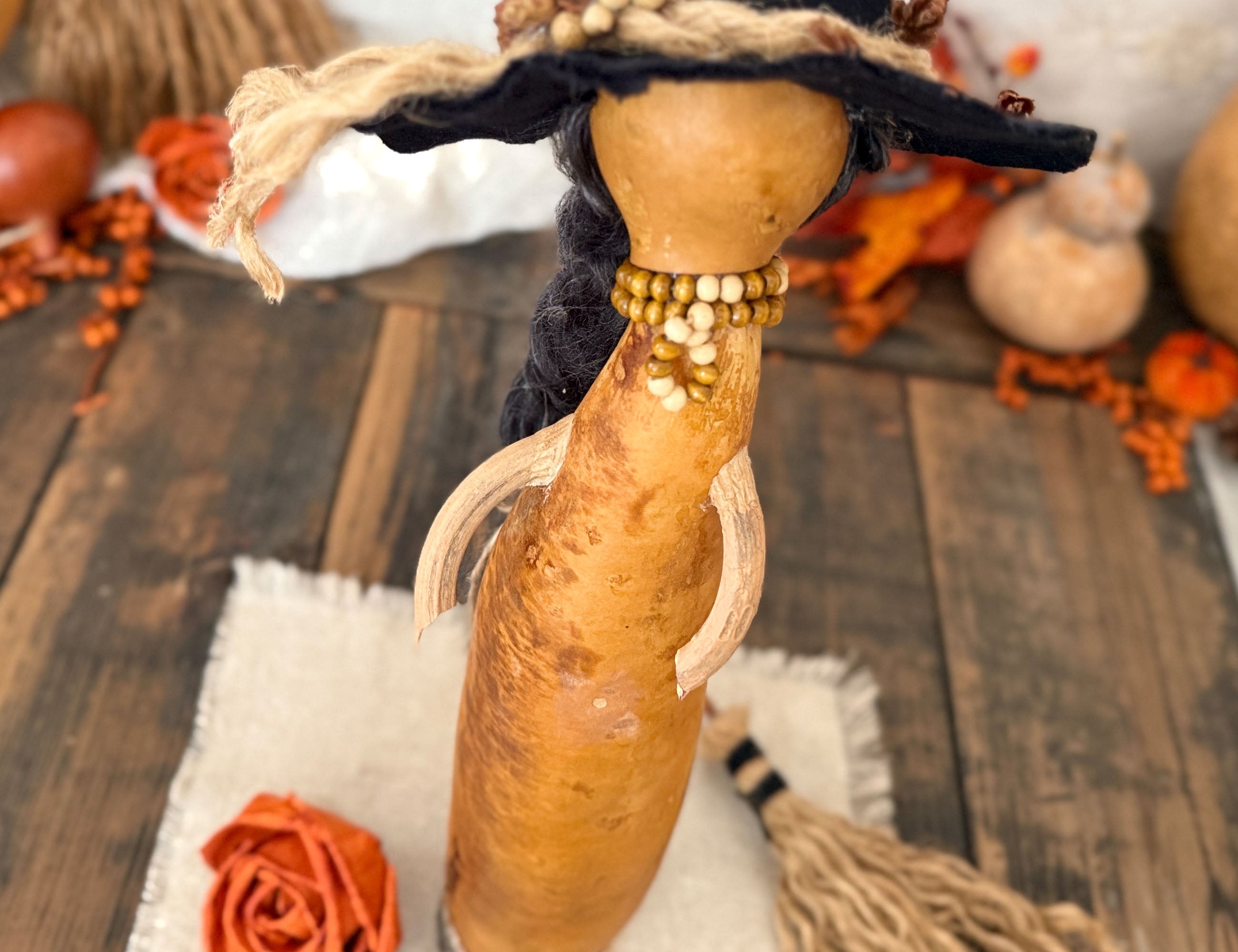 A handcrafted gourd doll with a black hat, braided black hair, and a beaded neck, displayed on a rustic wooden surface with autumn decorations.