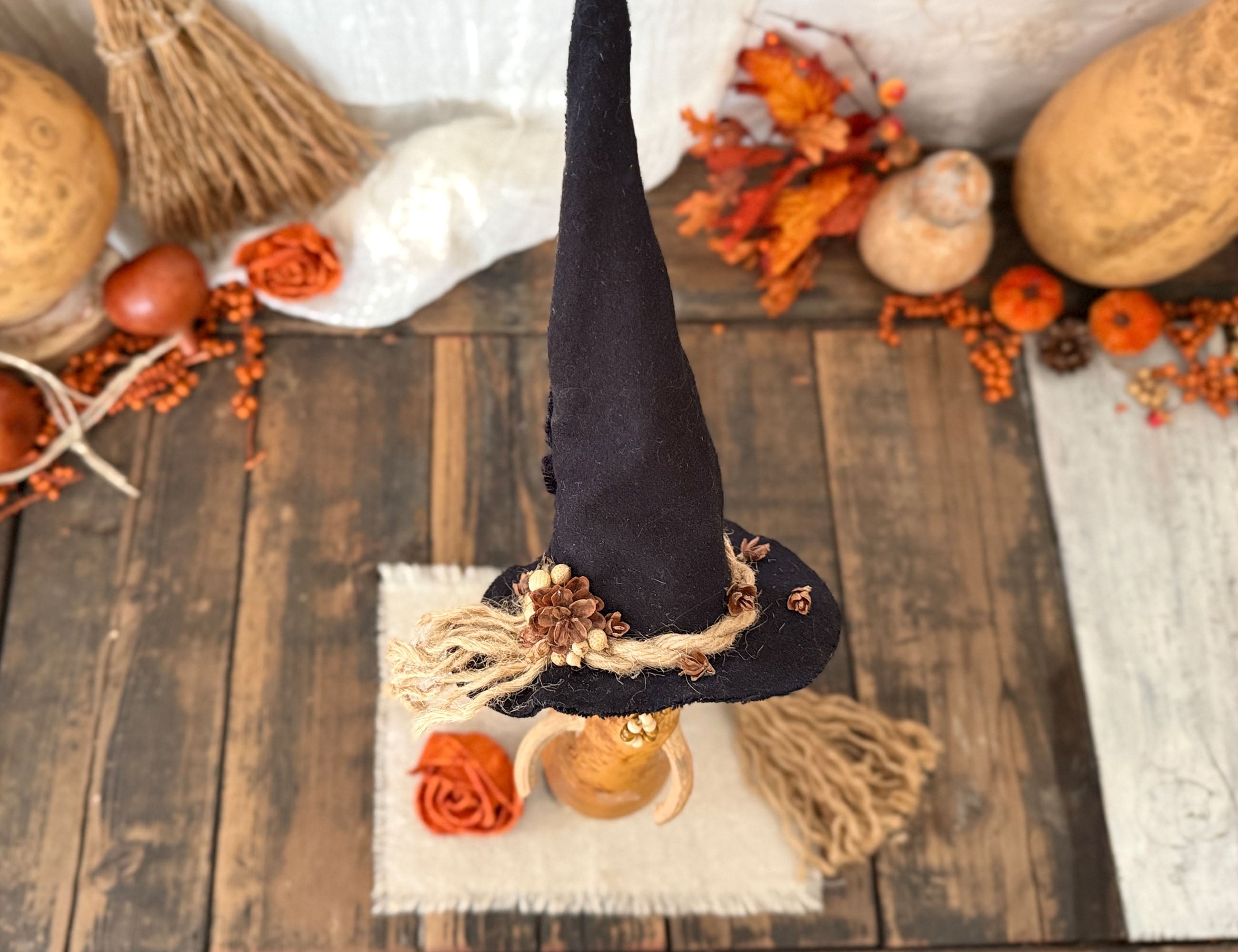 A top down view of a black witch hat on a gourd decoration with a burlap trim and small brown decorative accents, displayed on a rustic wooden surface with autumn props including pumpkins, orange berries, dried leaves, and a small candle.
