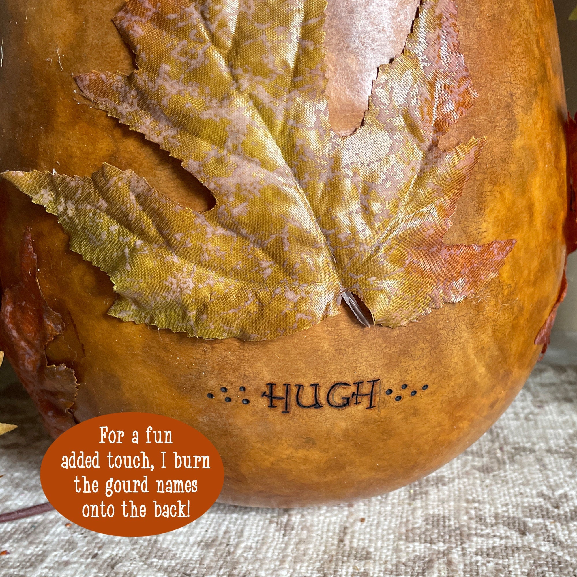 Gourd with a leaf decoration and 'HUGH' engraved, on a textured fabric background.