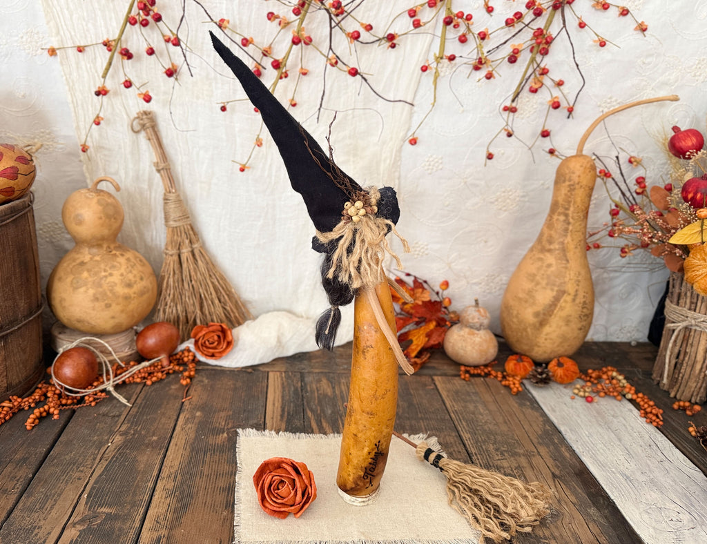 A decorative Halloween witch made from a hollowed gourd, featuring a tall black fabric hat, a black witch hat topper, and a beige jute tassel, displayed on a rustic wooden table surrounded by autumn decorations. Showing the right profile of the figurine.