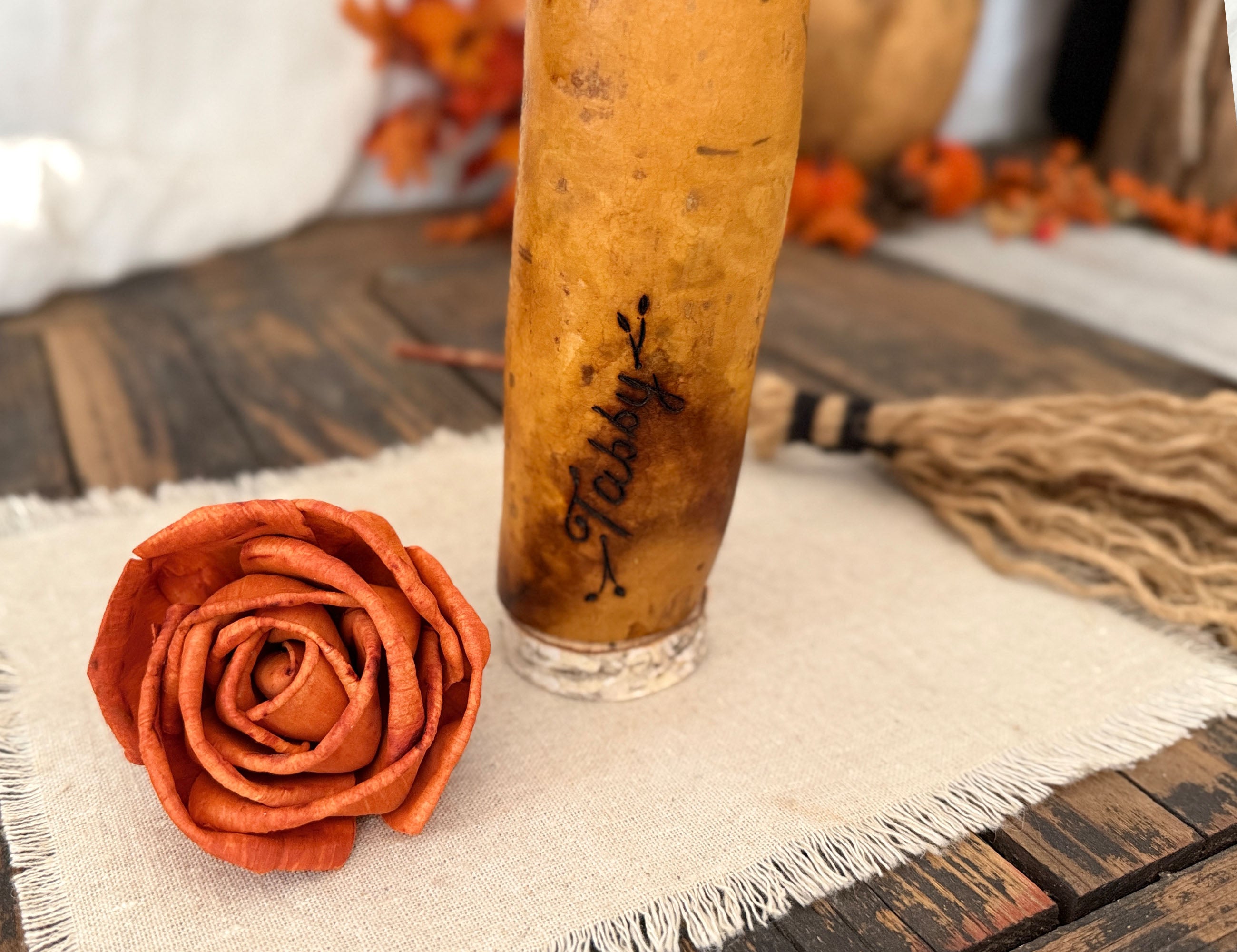 A closeup image showing this gourd witches name engraved on the lower left of her body. This witch is named "Tabby". A orange wood rose sits next to the gourd. 