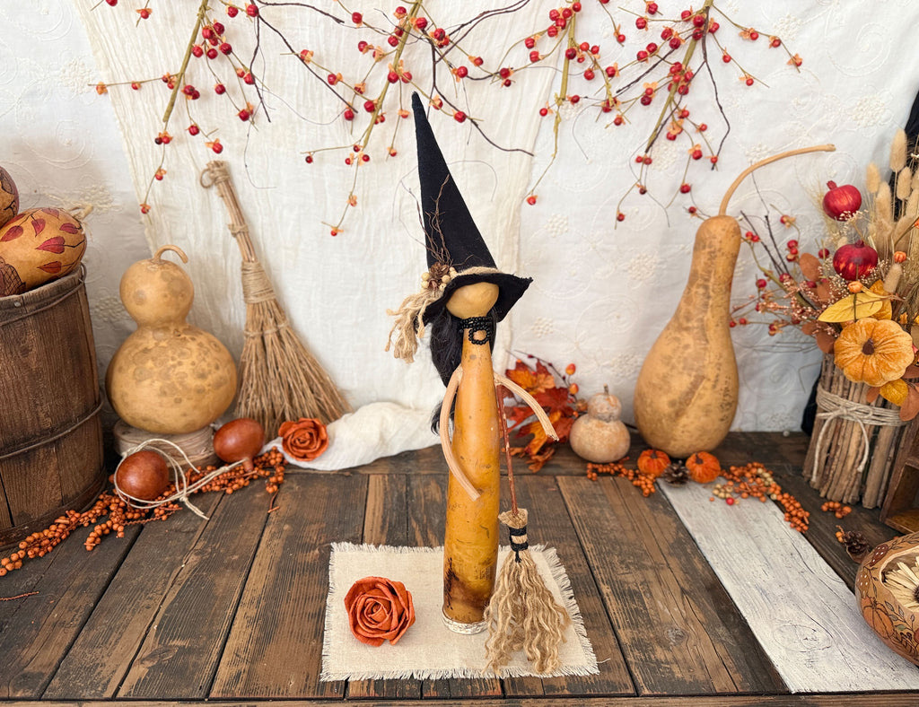 A decorative autumn scene with a central gourd figure wearing a pointed black hat and holding a broom, surrounded by gourds, berries, dried flowers, and rustic fall decor on a wooden surface against a white textured backdrop.