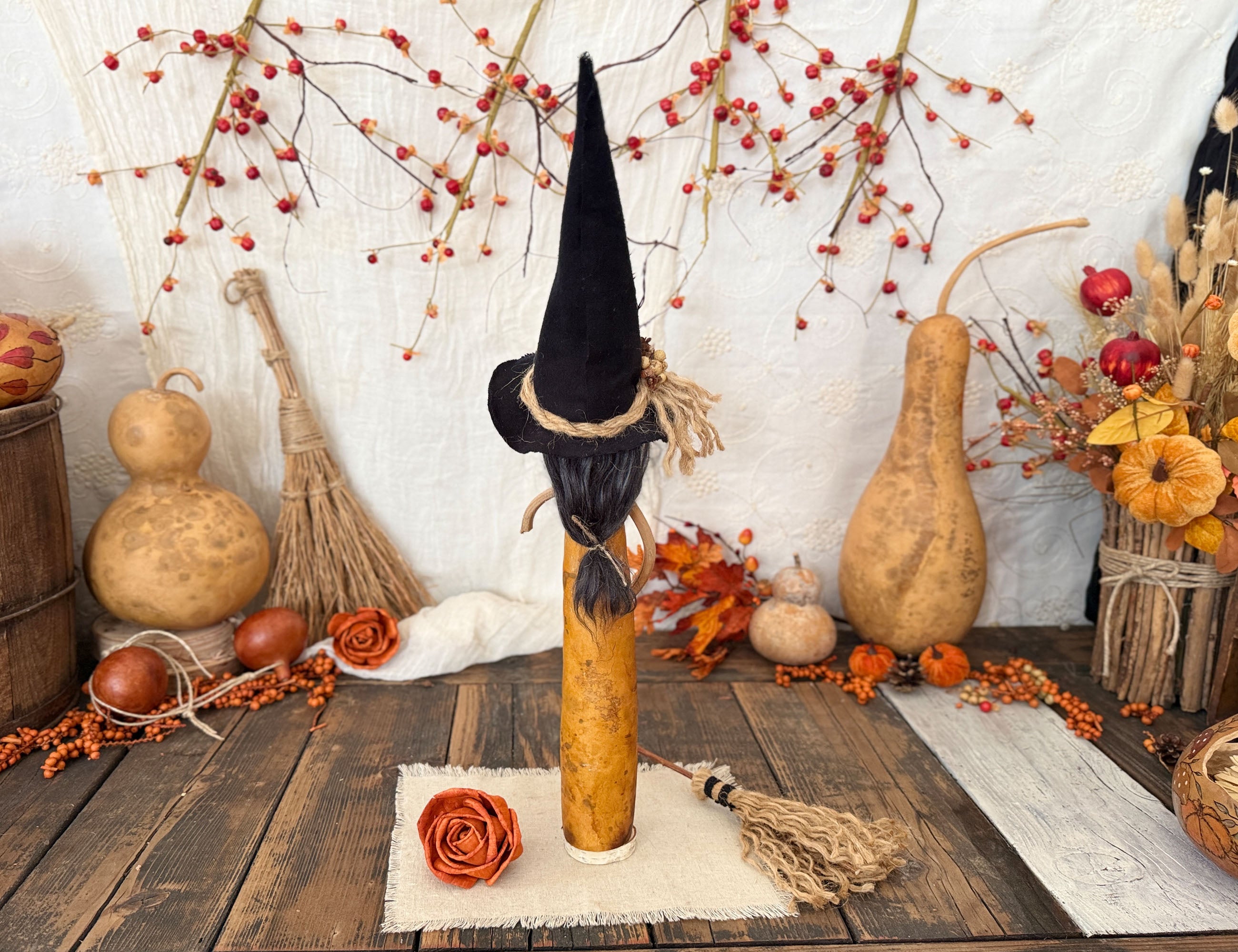 A Halloween decorative witch figure made from a yellow gourd with a black pointed hat and black hair, displayed on a rustic wooden table surrounded by autumn props including gourd sculptures, berries, dried flowers, and a small broom.
