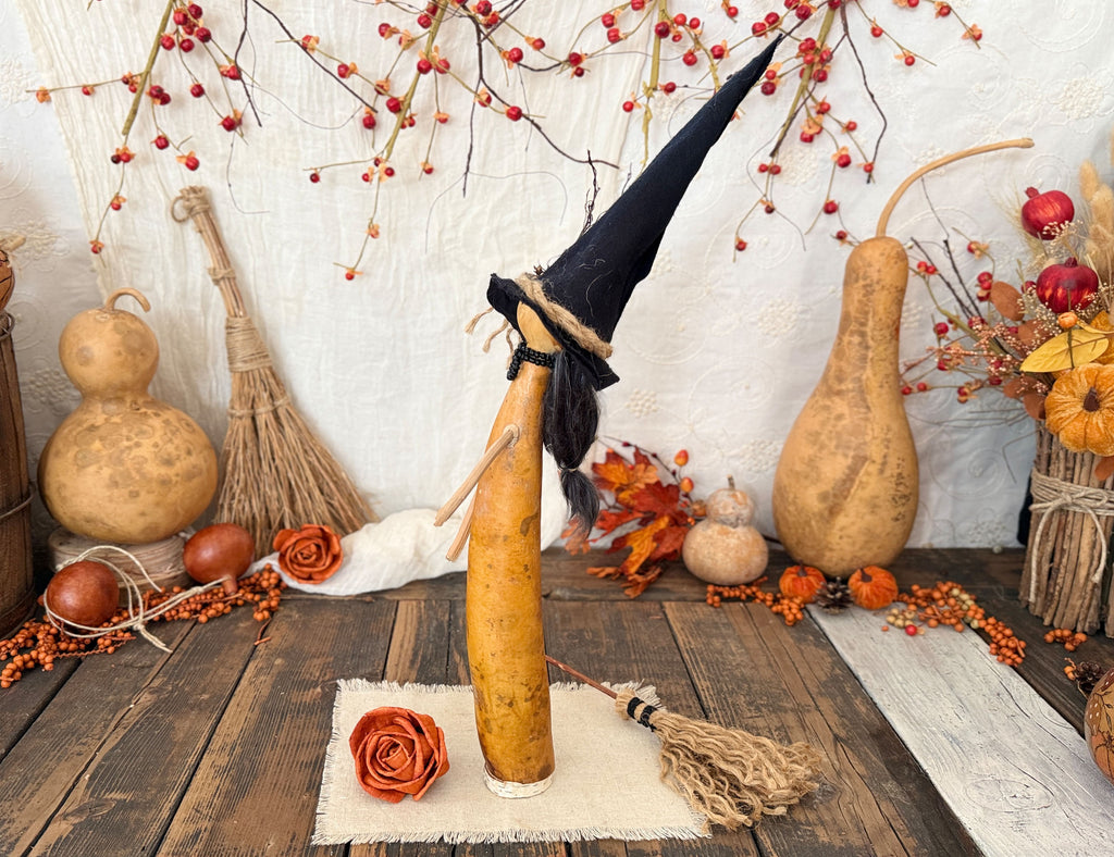 A decorative Halloween witch figure made from a gourd body with a tall black pointed hat, displayed on a rustic wooden surface with autumn props including gourds, berries, leaves, and a broom. Showing the left profile.