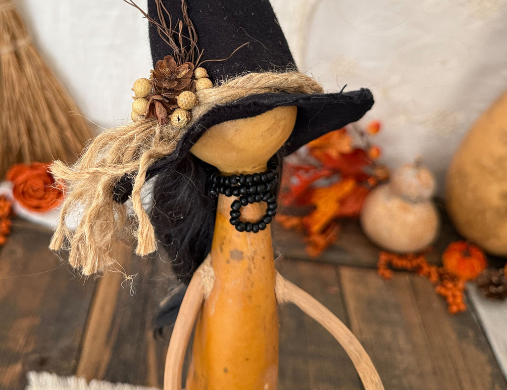 A rustic autumn decorative figure made from a gourd, wearing a black witch hat with jute trim and dried pinecones, standing on a wooden surface with fall decor in the background. The figurine does not have a face for a minimal witch aesthetic.