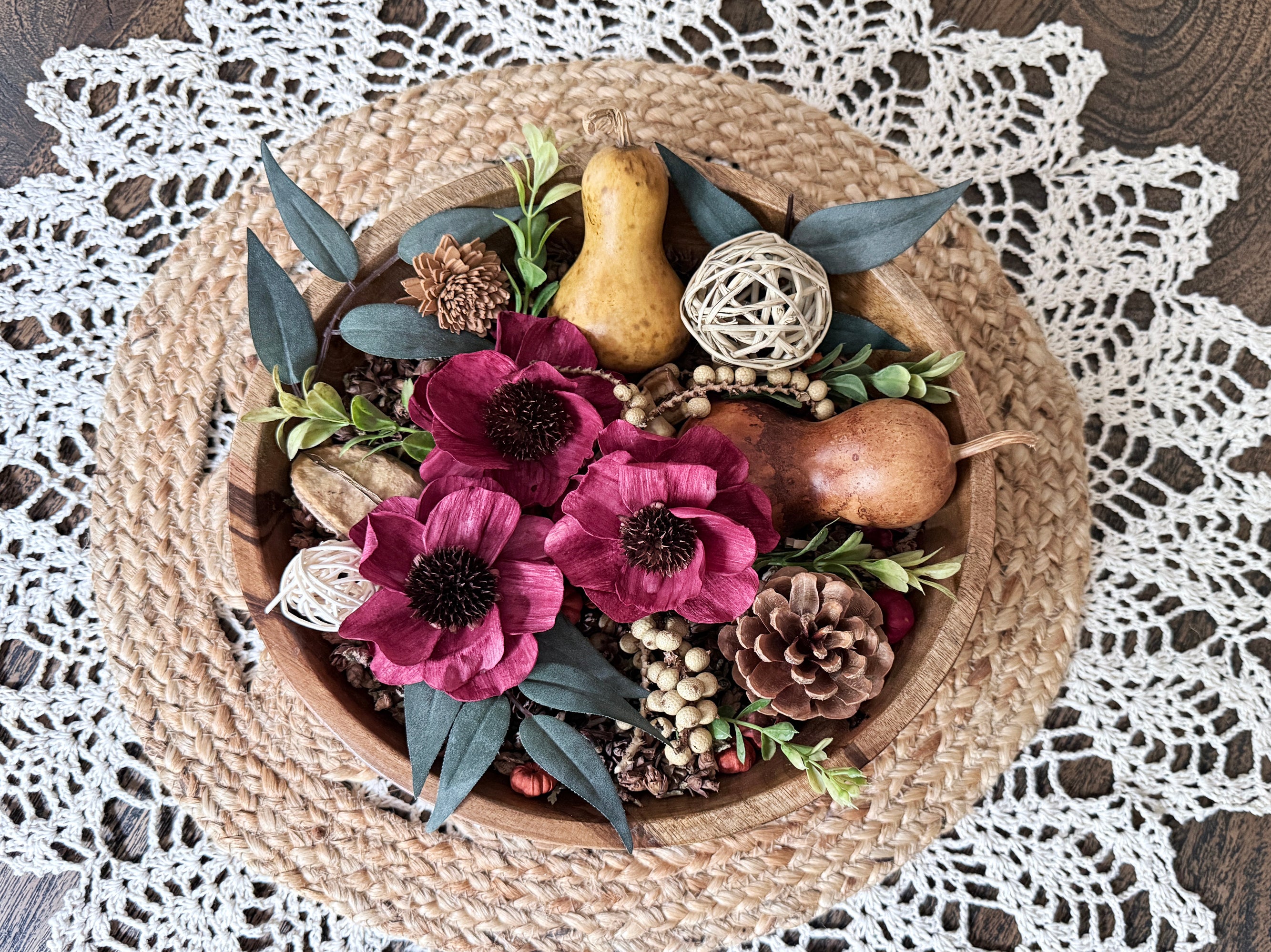 Decorative teak wood bowl with flowers, gourds, and pinecones on a lace tablecloth