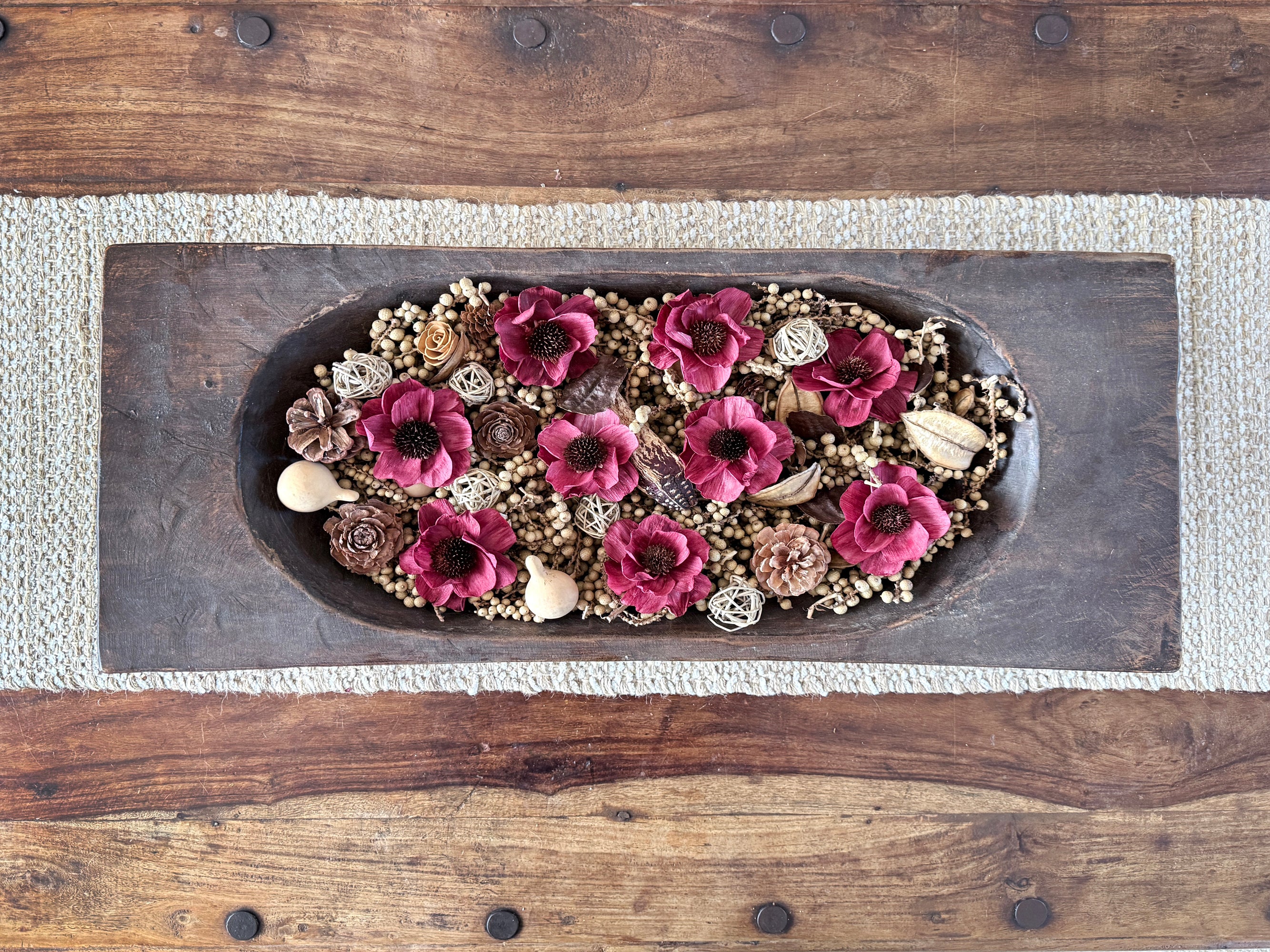 Decorative arrangement of red flowers and pinecones in a wooden dough bowl on a coffee table.