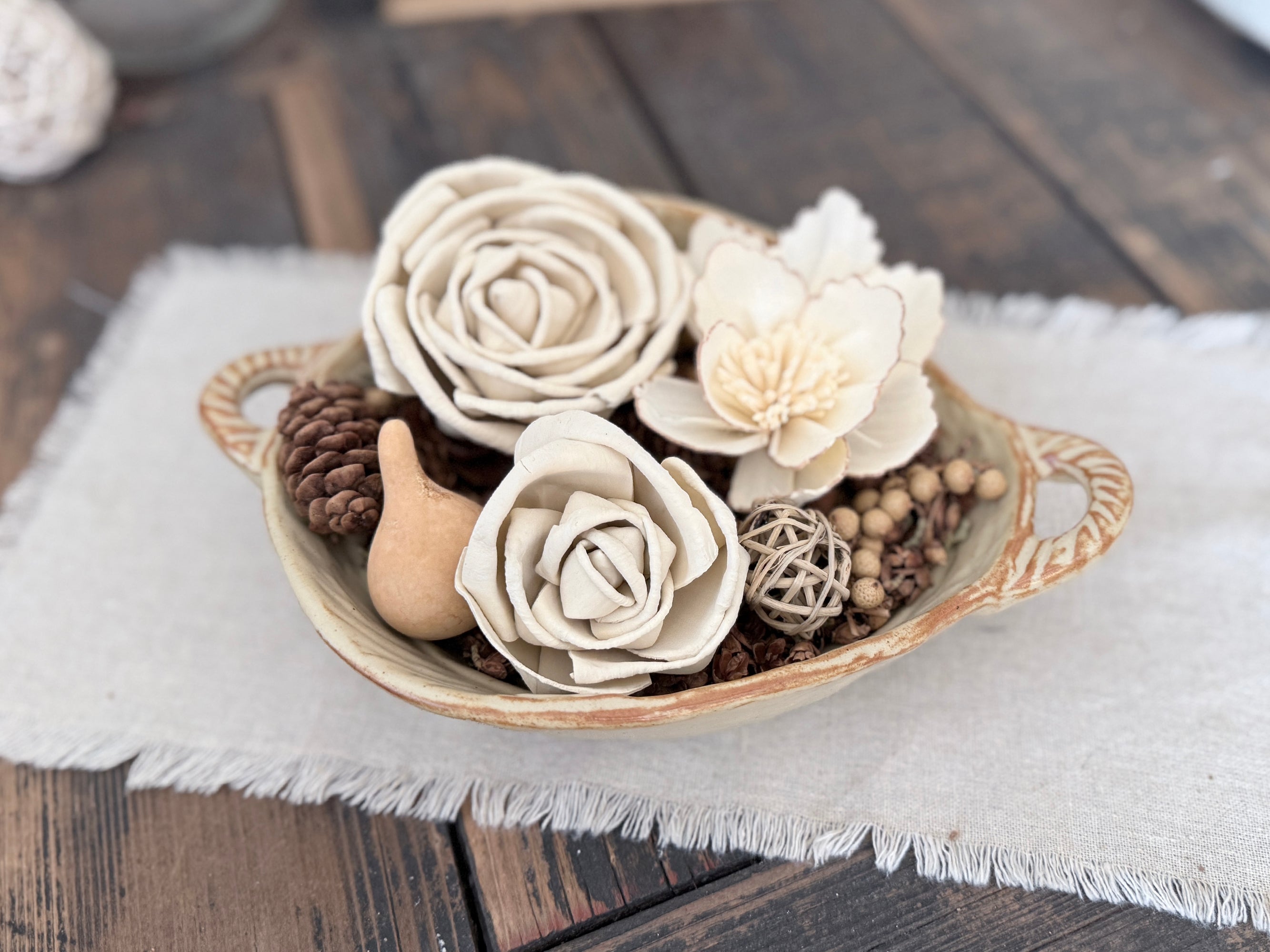 Decorative bowl with white wood flowers, pinecones, and a snail on a wooden surface.