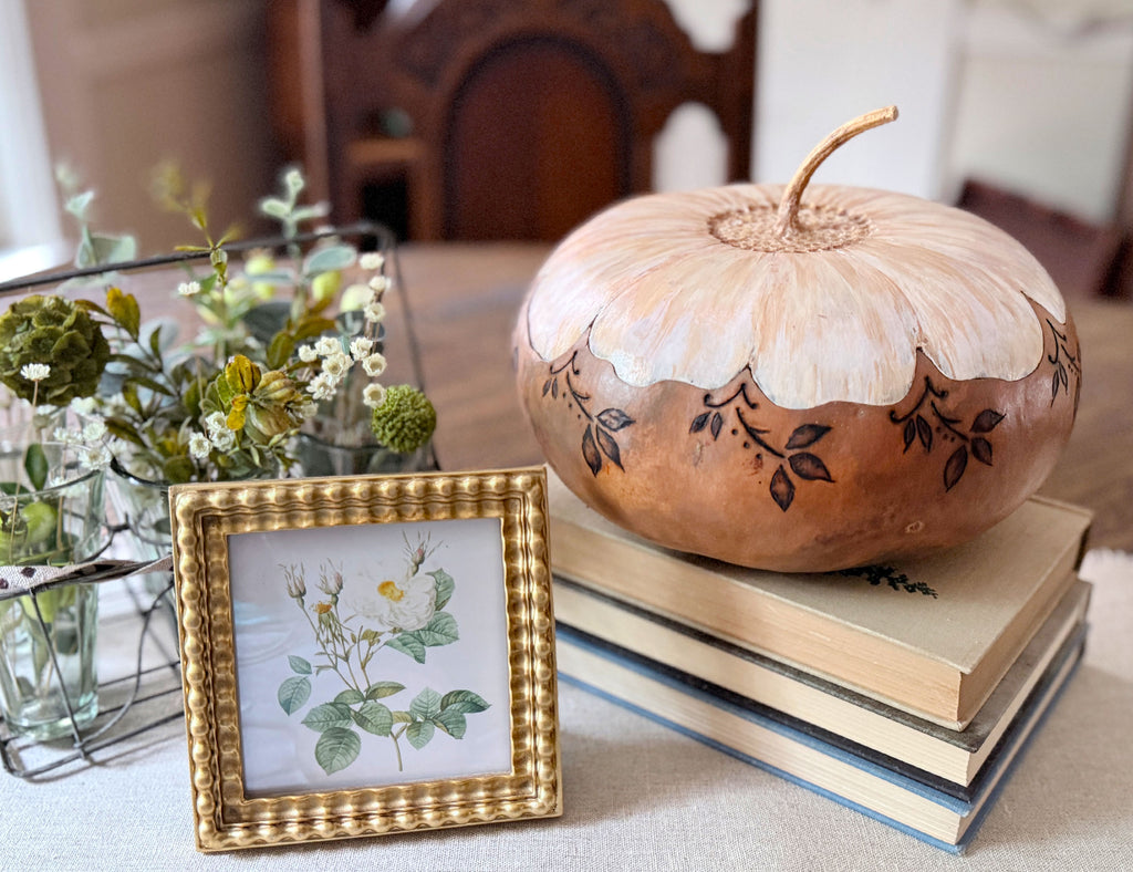 Artisan gourd vessel sitting on a stack of vintage books. The bottom half of the gourd is a neutral beige and the top has a fitted lid painted like a daisy. 