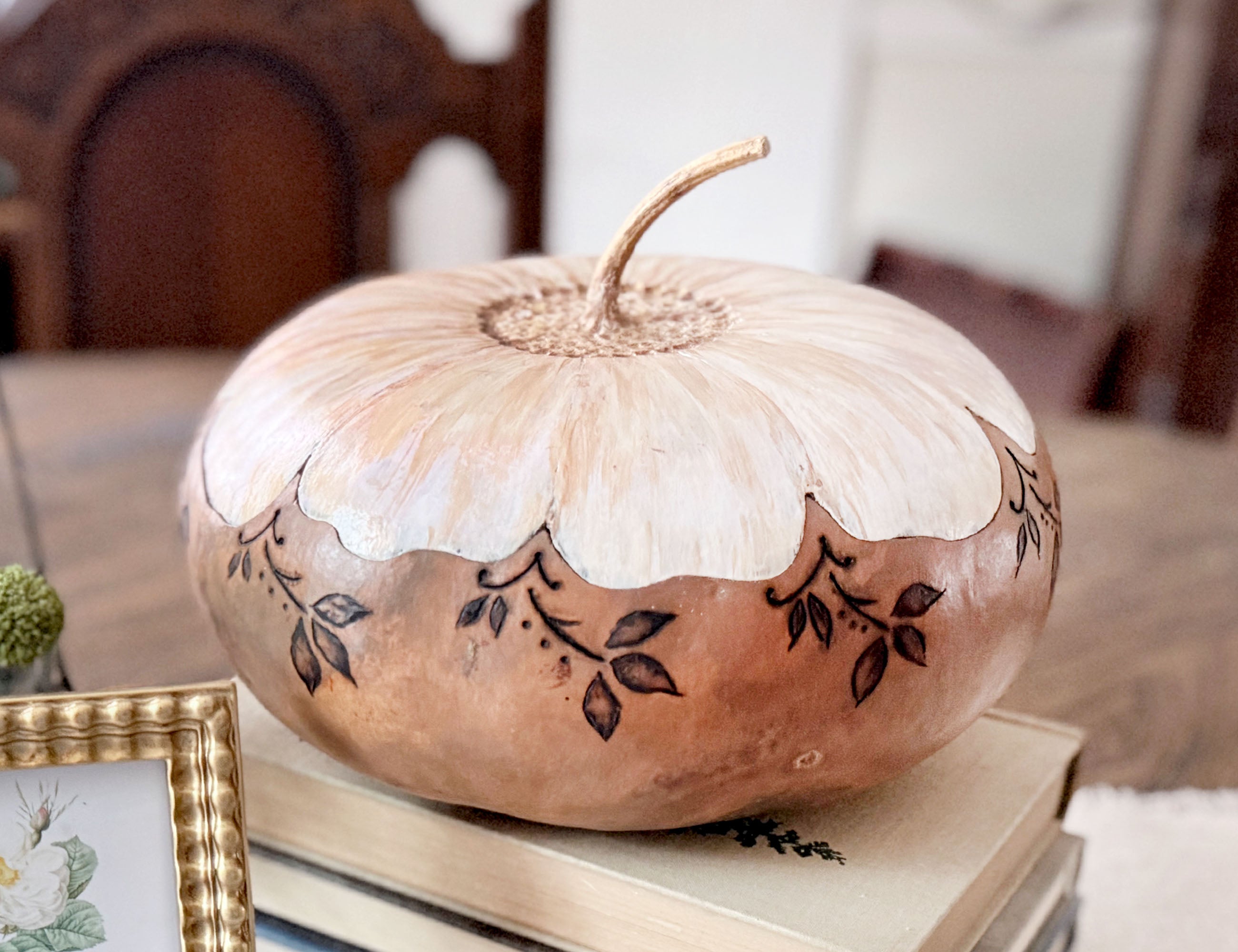Artisan gourd vessel sitting on a stack of vintage books. The bottom half of the gourd is a neutral beige and the top has a fitted lid painted like a daisy. 
