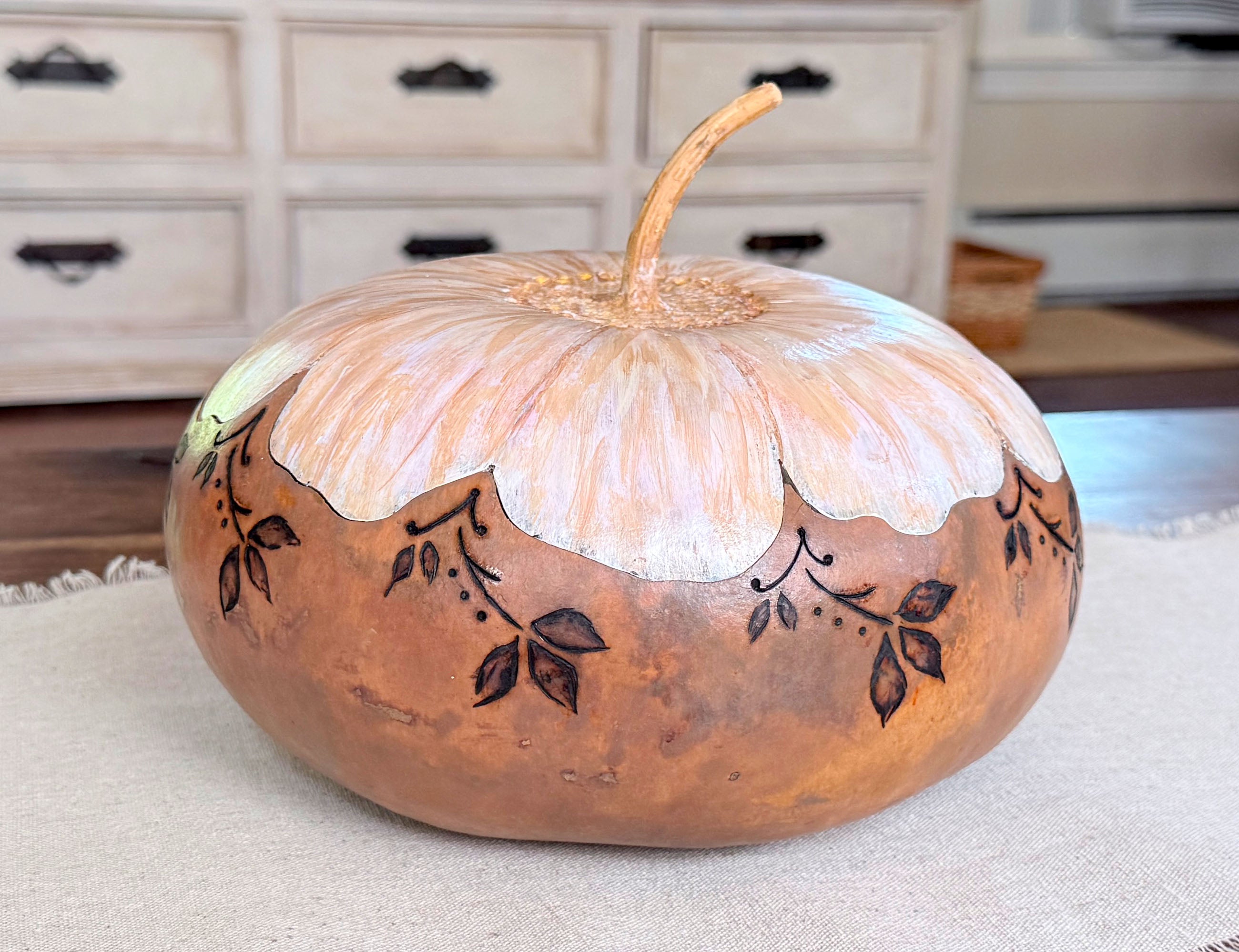 Artisan gourd vessel sitting on a wooden coffee table. The bottom half of the gourd is a neutral beige and the top has a fitted lid painted like a daisy. 