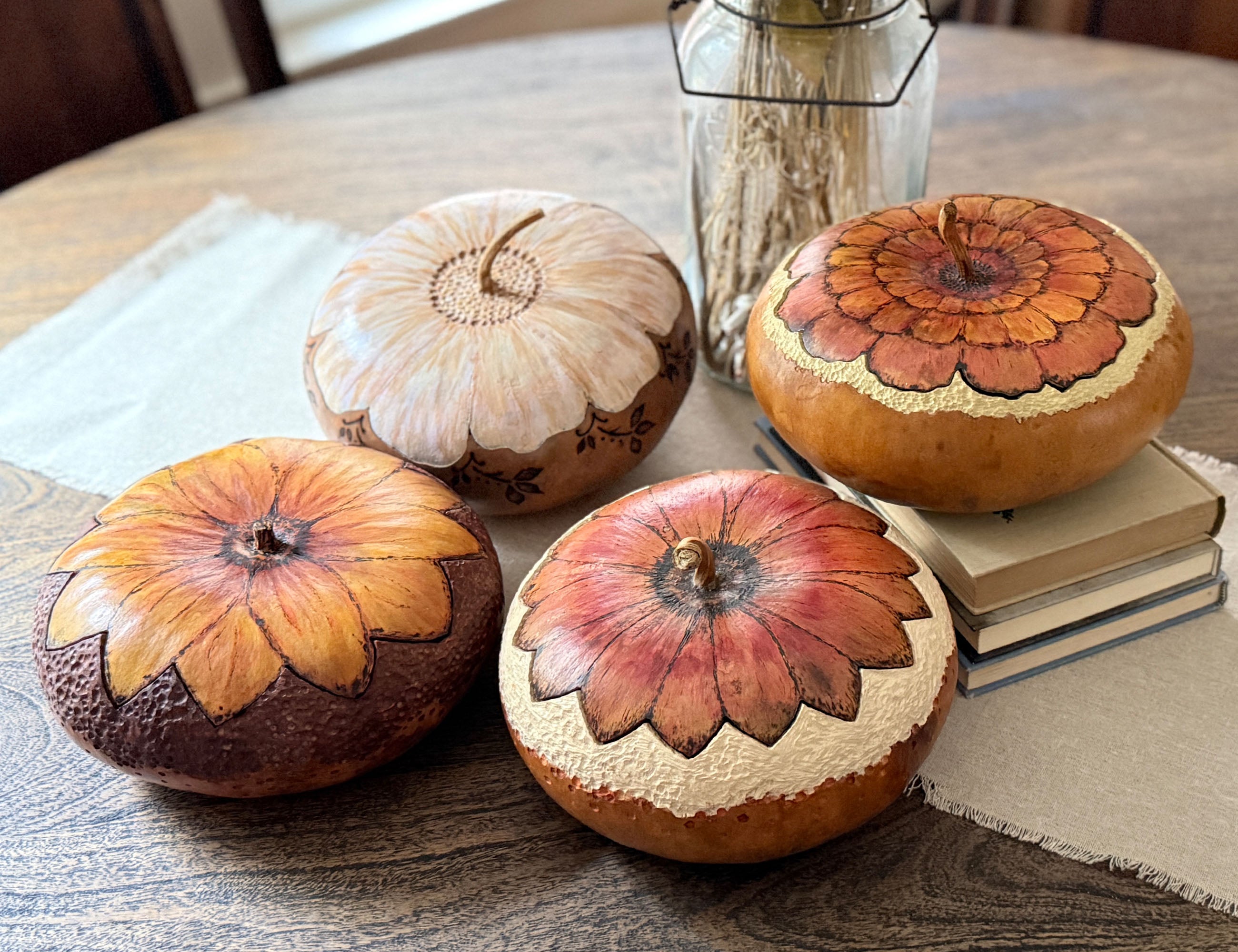 Four decorative hand-painted gourd bowls with fitted flower blossom lids arranged on a wooden table with a book and a glass jar in the background.