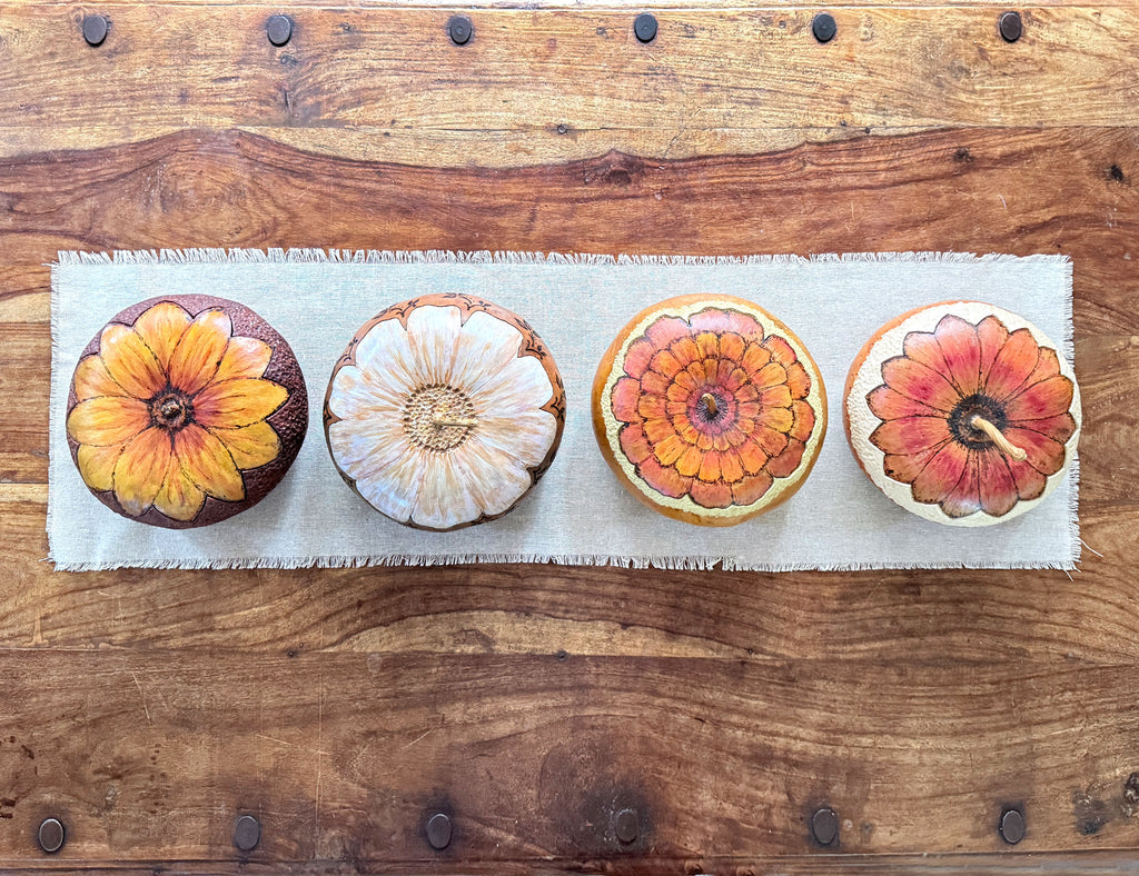 Four decorative gourd bowls arranged in a row on a light beige fabric runner over a rustic wooden coffee table. The gourds have painted floral lids  in autumn colors including orange, yellow, red, and white.