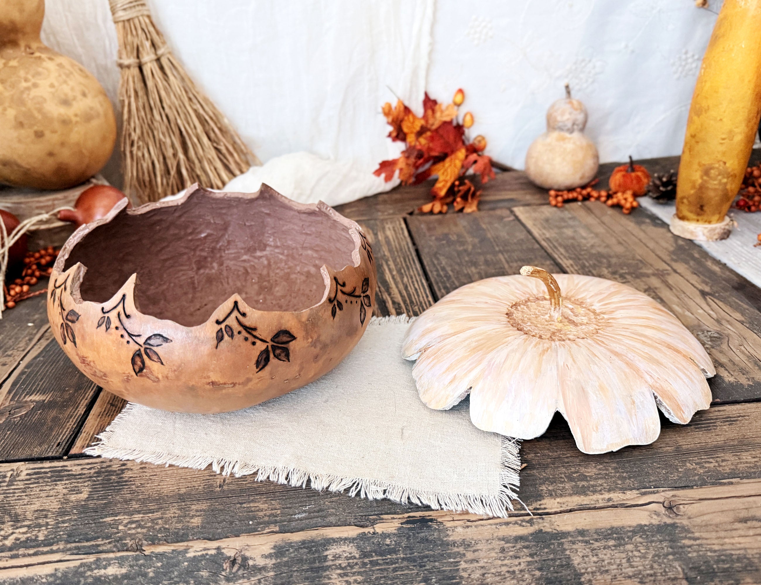 A decorative gourd bowl with the lid sitting next to it. Bowl is neutral beige with wood burned leaves around the rim. The flower blossom shape lid sits next to it.