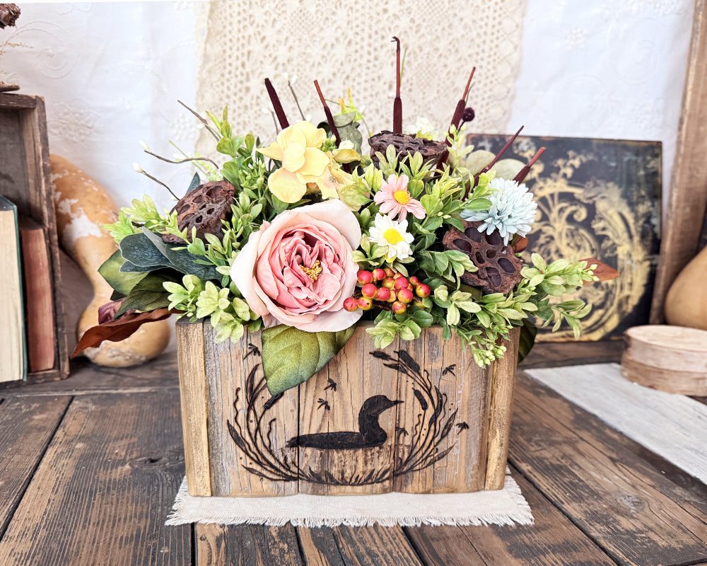 Floral arrangement in a handcrafted wooden box with a wood burned loon design on a rustic wooden surface.