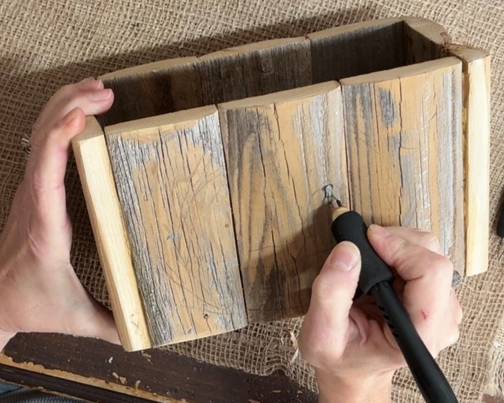 Person using a pyrography pen to engrave  to a wooden box on a textured surface.