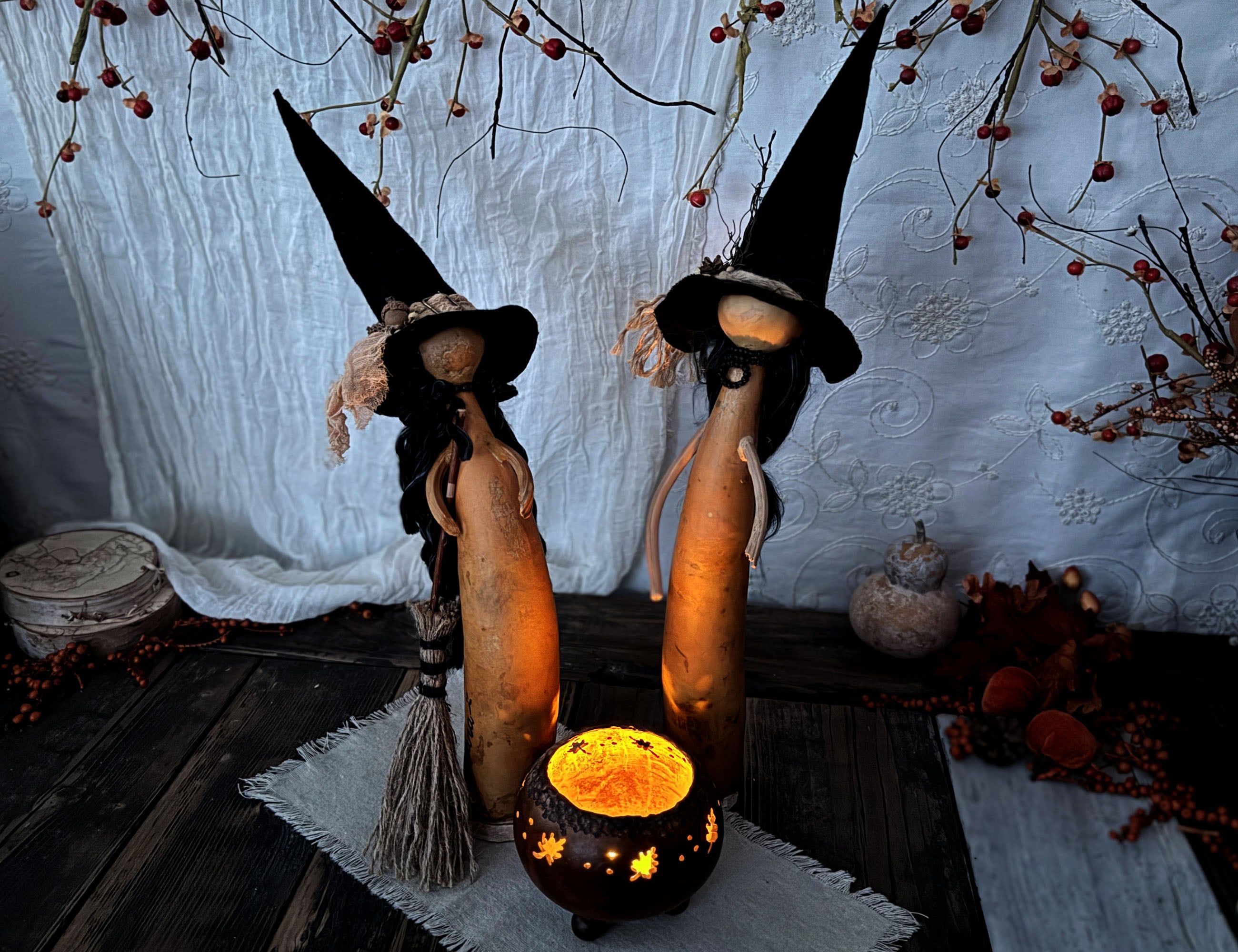 Two decorative witch figurines made from hollowed gourds, wearing tall black pointed hats and standing on a rustic wooden surface in front of a white fabric backdrop with dried berries and autumn decor.
