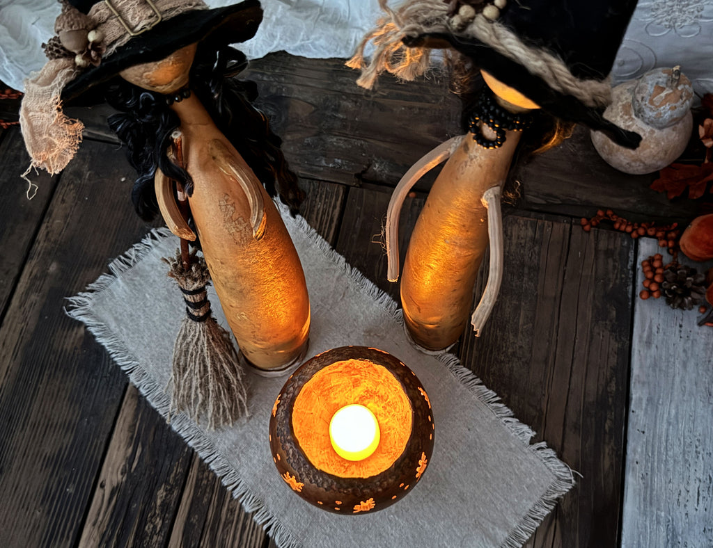 A styled tabletop scene with two witch-themed gourd decorations wearing black hats and standing beside a small glowing cauldron holder on a gray cloth over a dark wood surface.