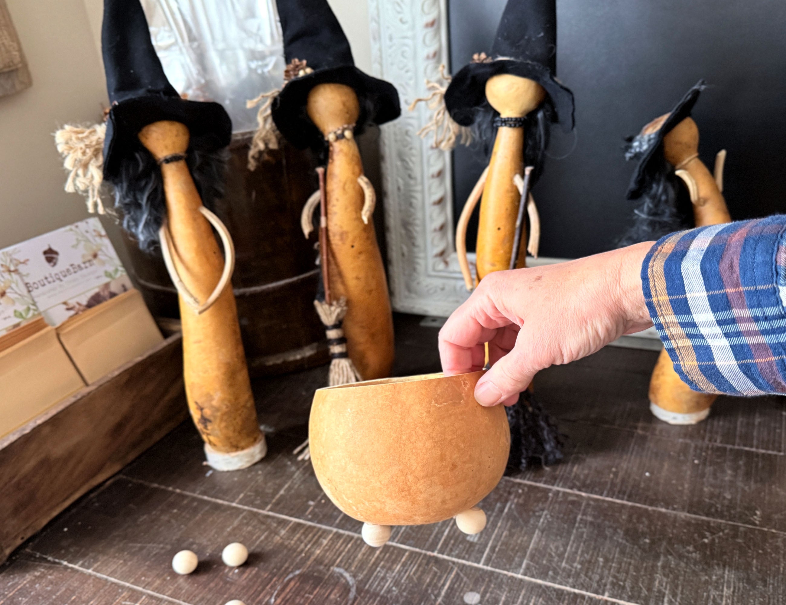 A hand holds a small carved gourd bowl with three legs in a rustic indoor setting, with several witch figurines wearing black hats in the background.