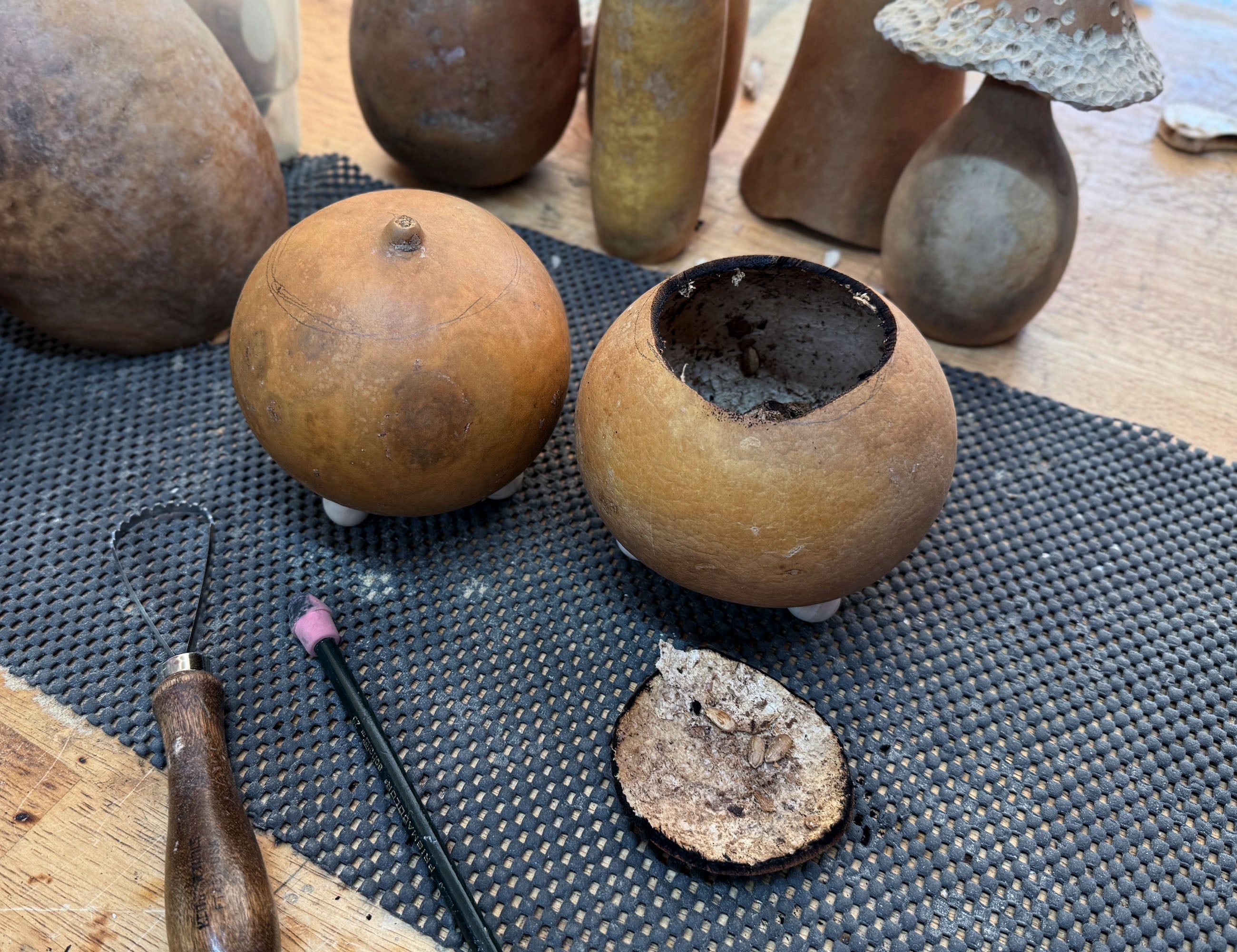 Two round gourds  one whole and one hollowed out, displayed on a black textured mat with sculpting tools and other clay vessels in the background.