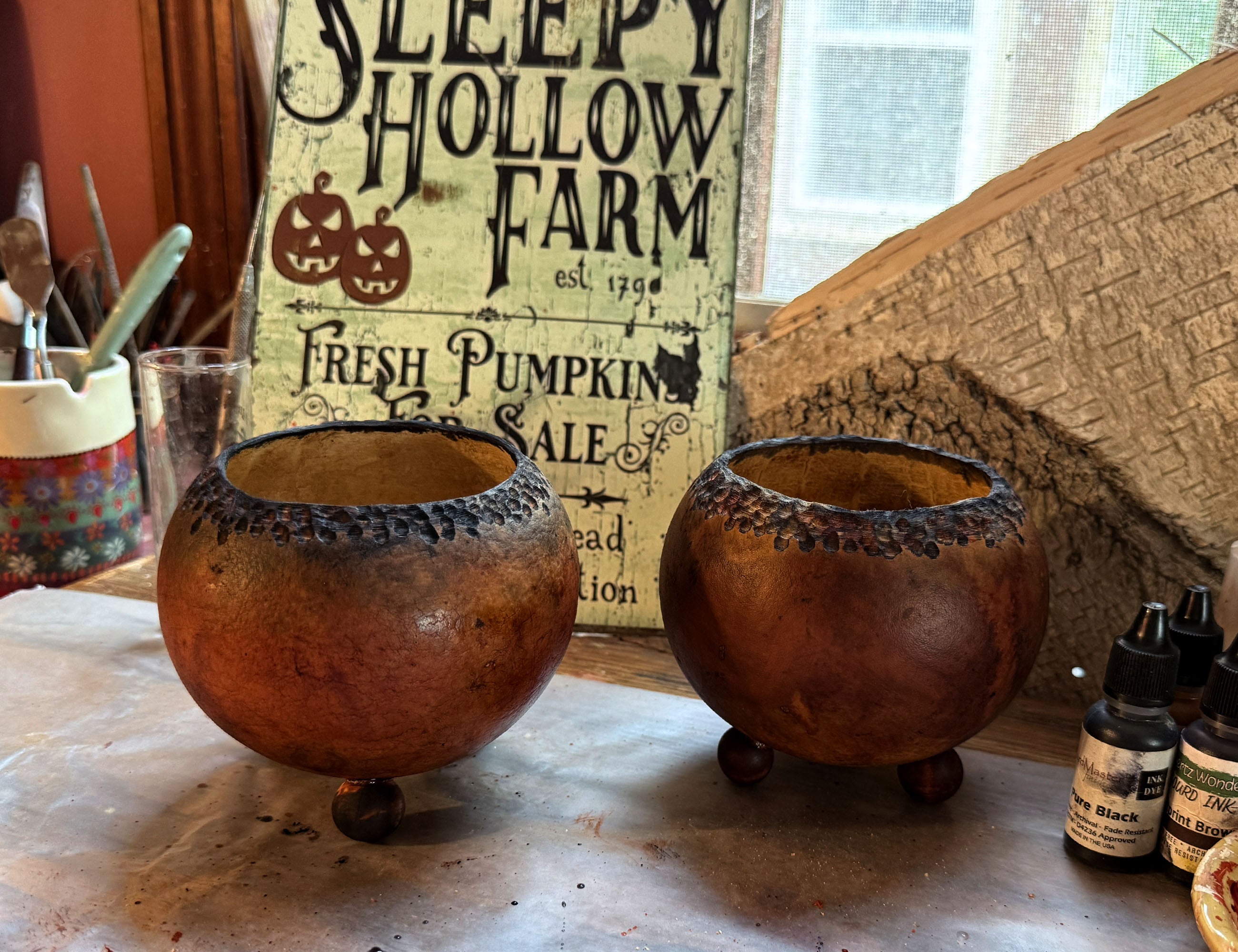 Two decorative dried gourd footed cauldrons displayed on a tabletop in a rustic indoor setting, with a vintage 'Sleepy Hollow Farm' sign and Halloween-themed bottles in the background.