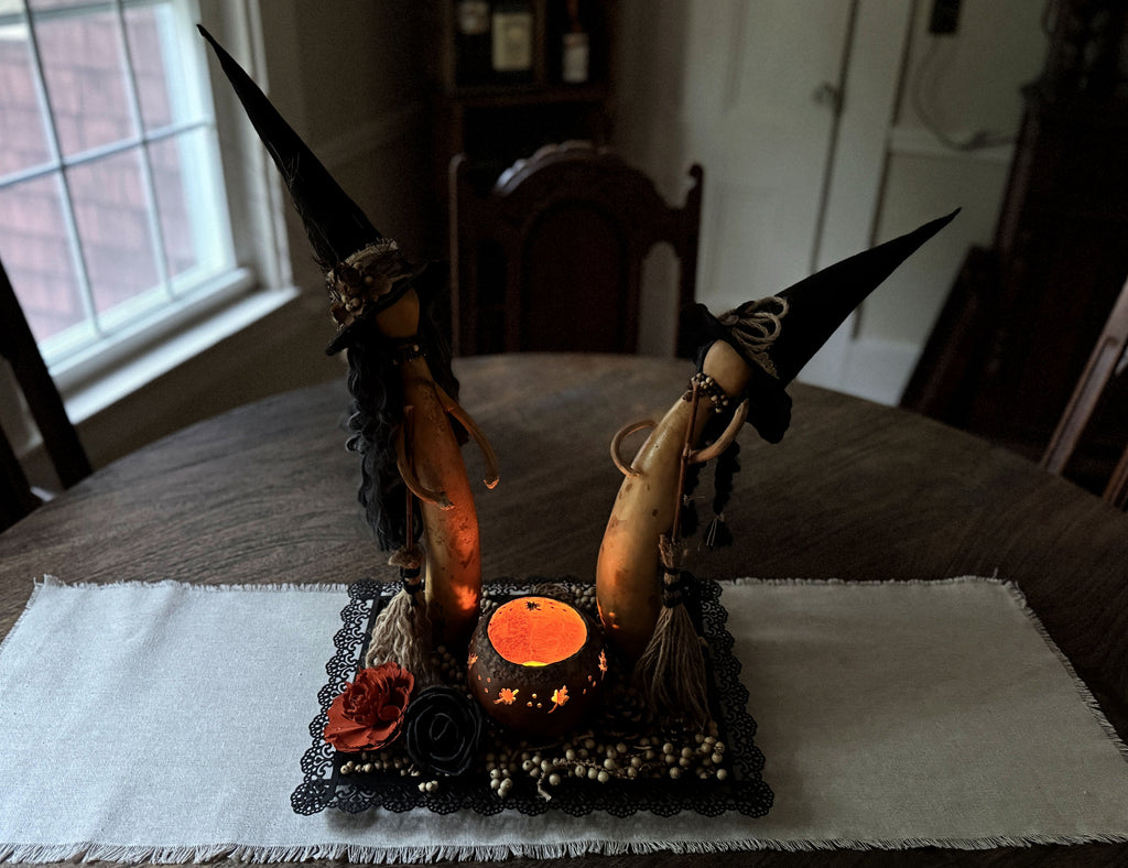 A Halloween-themed tabletop centerpiece featuring two witch figurines with tall black hats, standing on a black lace tray with a glowing orange cauldron and decorative accents, set on a white placemat on a wooden table.