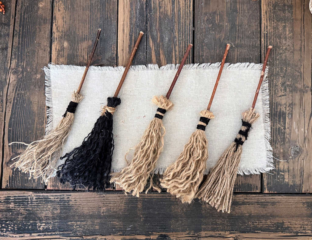 Four miniature broomsticks with jute-tassel heads are arranged on a white fringed fabric runner over a dark rustic wooden surface. Three brooms have beige/tan tassels and one has a black tassel.