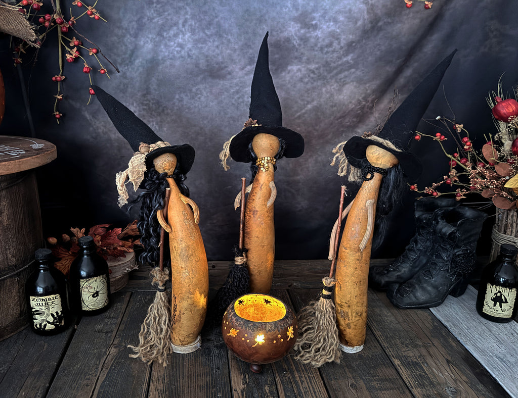 Three rustic witch figurines stand around a gourd cauldron lit with a candle. A spooky nigh sky is in the back with fall berries and leaves.