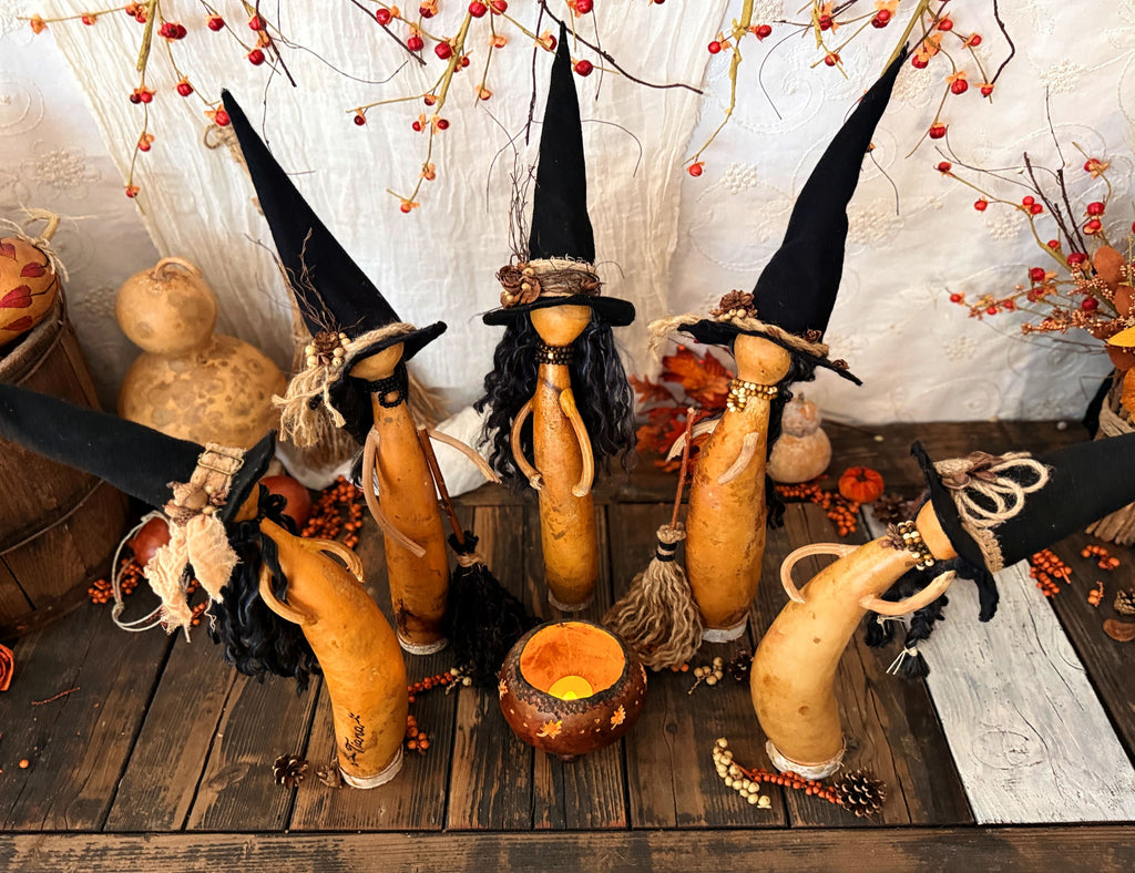 Halloween decorative witch figurines crafted from gourds and gourd stems are arms, wearing tall black pointed hats, arranged on a rustic wooden surface with a small cauldron lantern and autumn berries.