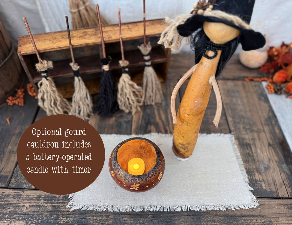 A rustic Halloween decoration set on a wooden table, featuring a gourd cauldron with a lit battery-operated candle, a witch figure made from a gourd, a wooden broom rack with several small brooms, and a brown text bubble describing the battery-operated candle option.