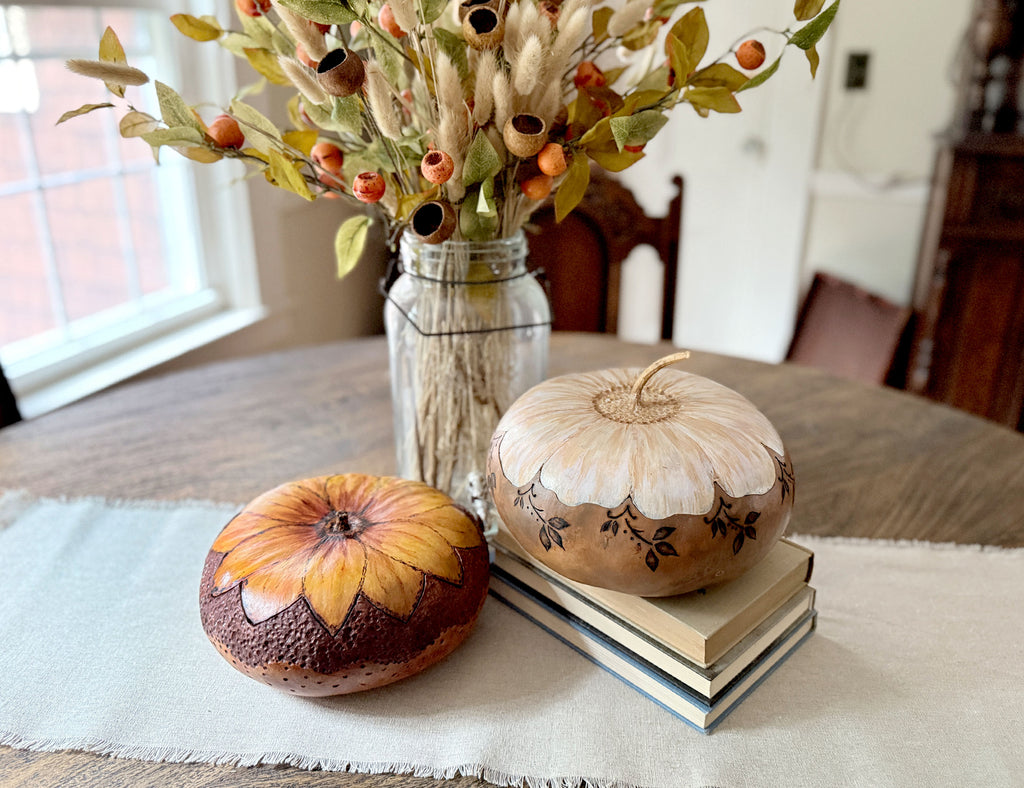 Two decorative gourd bowls with lids styled on a table runner in a dining room setting. One gourd is painted with a yellow and orange sunflower pattern on a dark brown base, while the other is cream-colored with hand-painted black leaf details and rests on a stack of books.