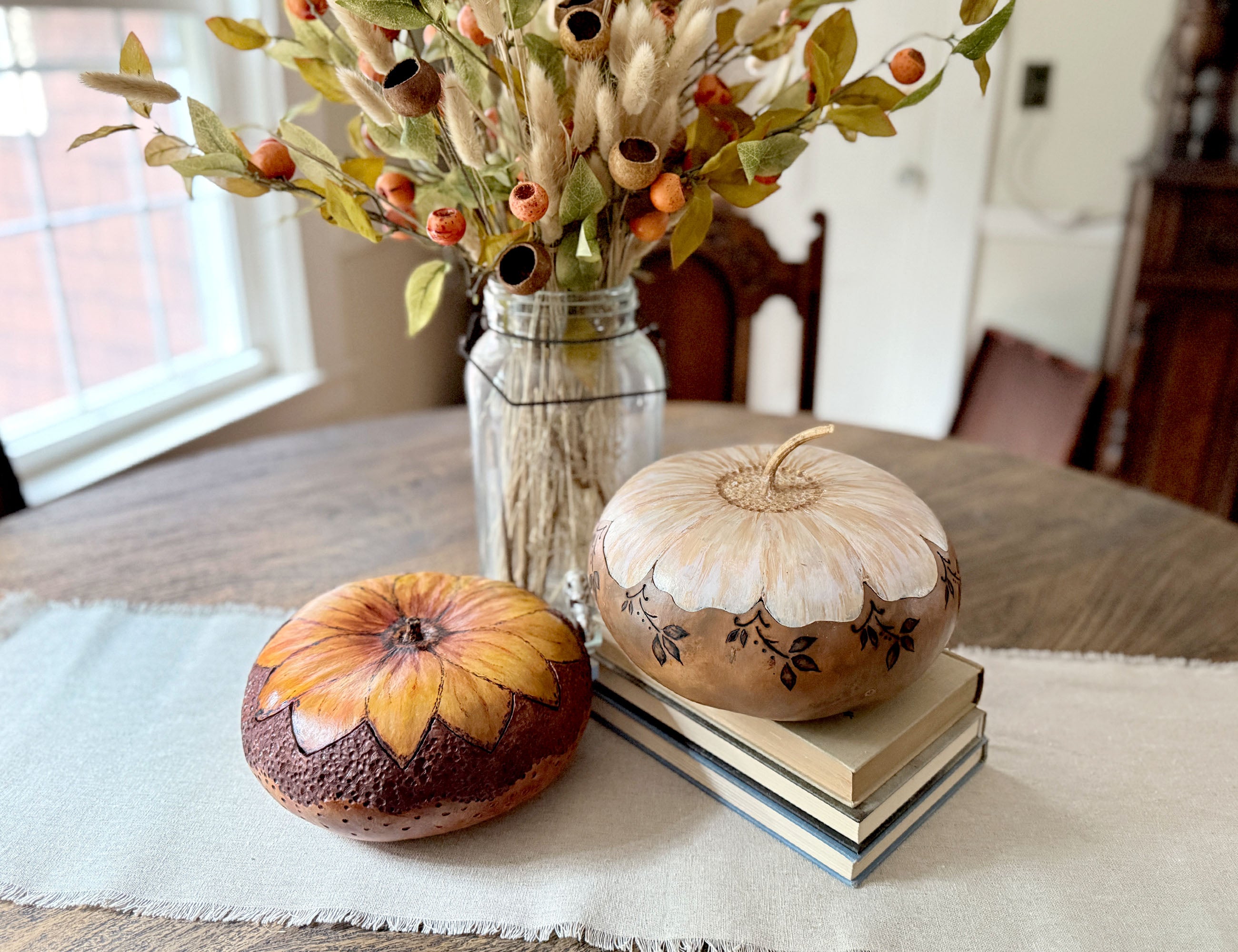 Two decorative gourd bowls with lids styled on a table runner in a dining room setting. One gourd is painted with a yellow and orange sunflower pattern on a dark brown base, while the other is cream-colored with hand-painted black leaf details and rests on a stack of books.