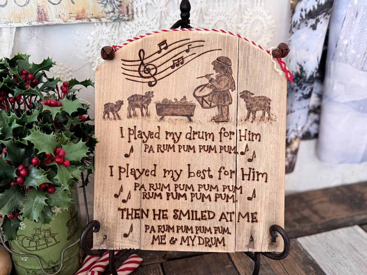 A wood wall sign made from upcycled wood with a rounded top. Artwork features the Little Drummer Boy standing near the baby Jesus with sheep near. Musical notes are at the top, with words from the song lyrics below. The artwork has  been wood burned onto the wood.  The top has red and white cord attached with rusty jingle bells. A bouquet of holly berries sits to the left. 