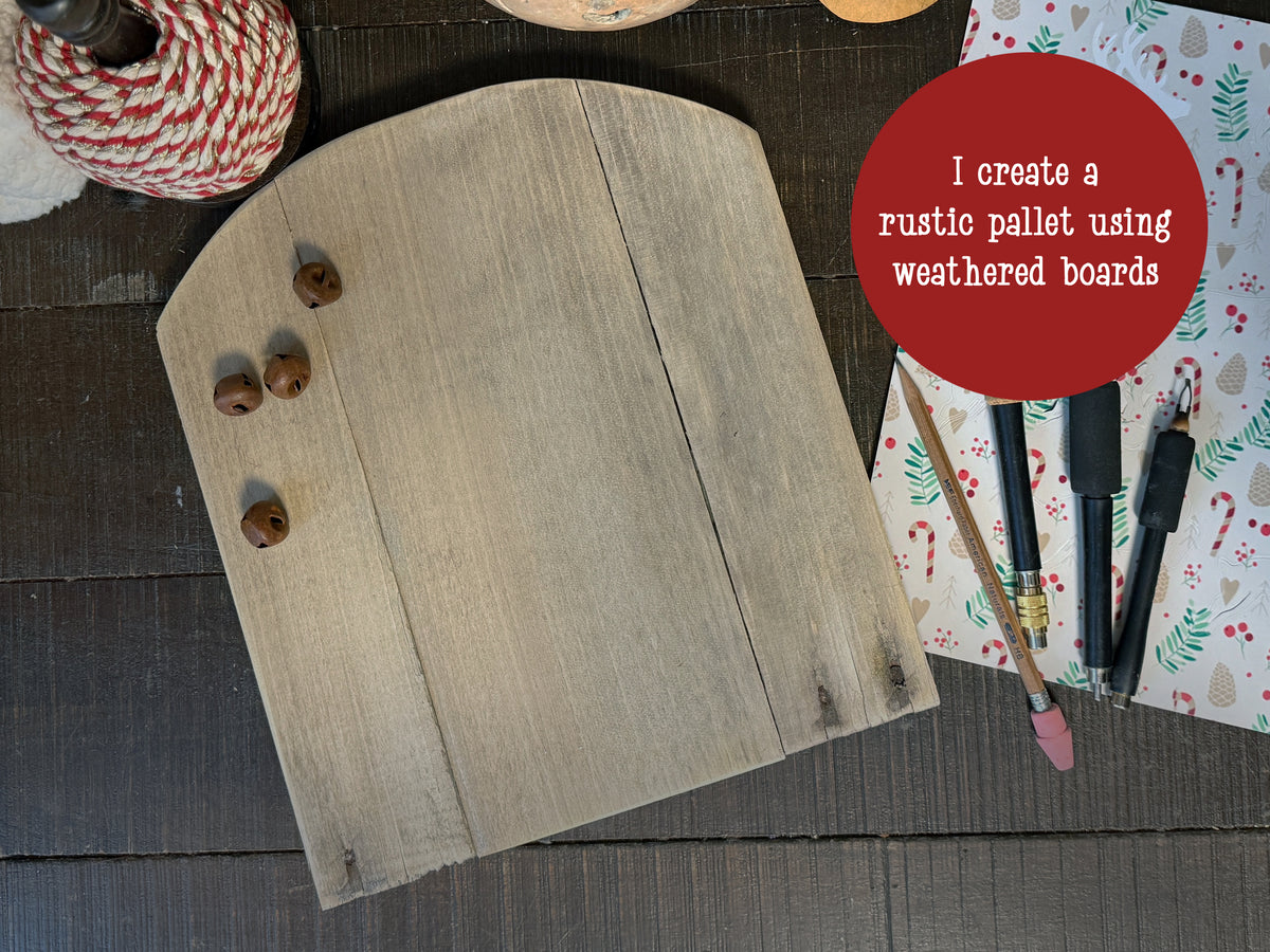 A wood pallet with a curved top is laid out on a table. Red and white cord sits near, rusty jingle bells lay on the wood. Pyrography pens lay near by with a pencil.