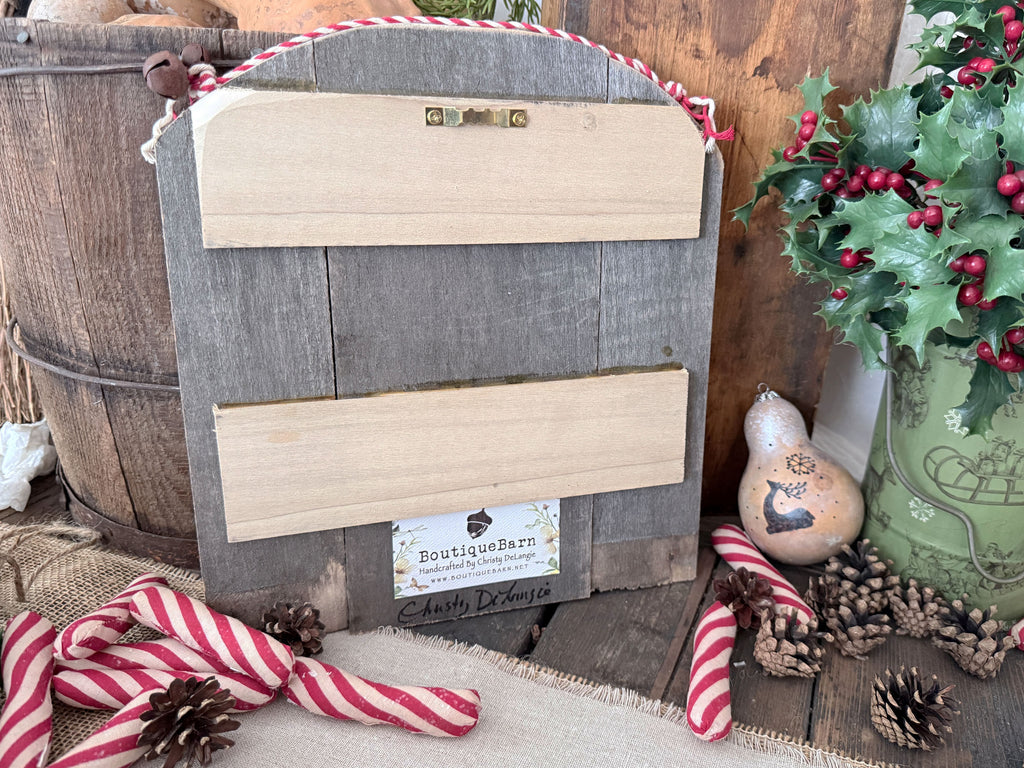Image shows the back of the Christmas sign with a saw tooth hanger attached at the top. Artist signature is at the bottom.