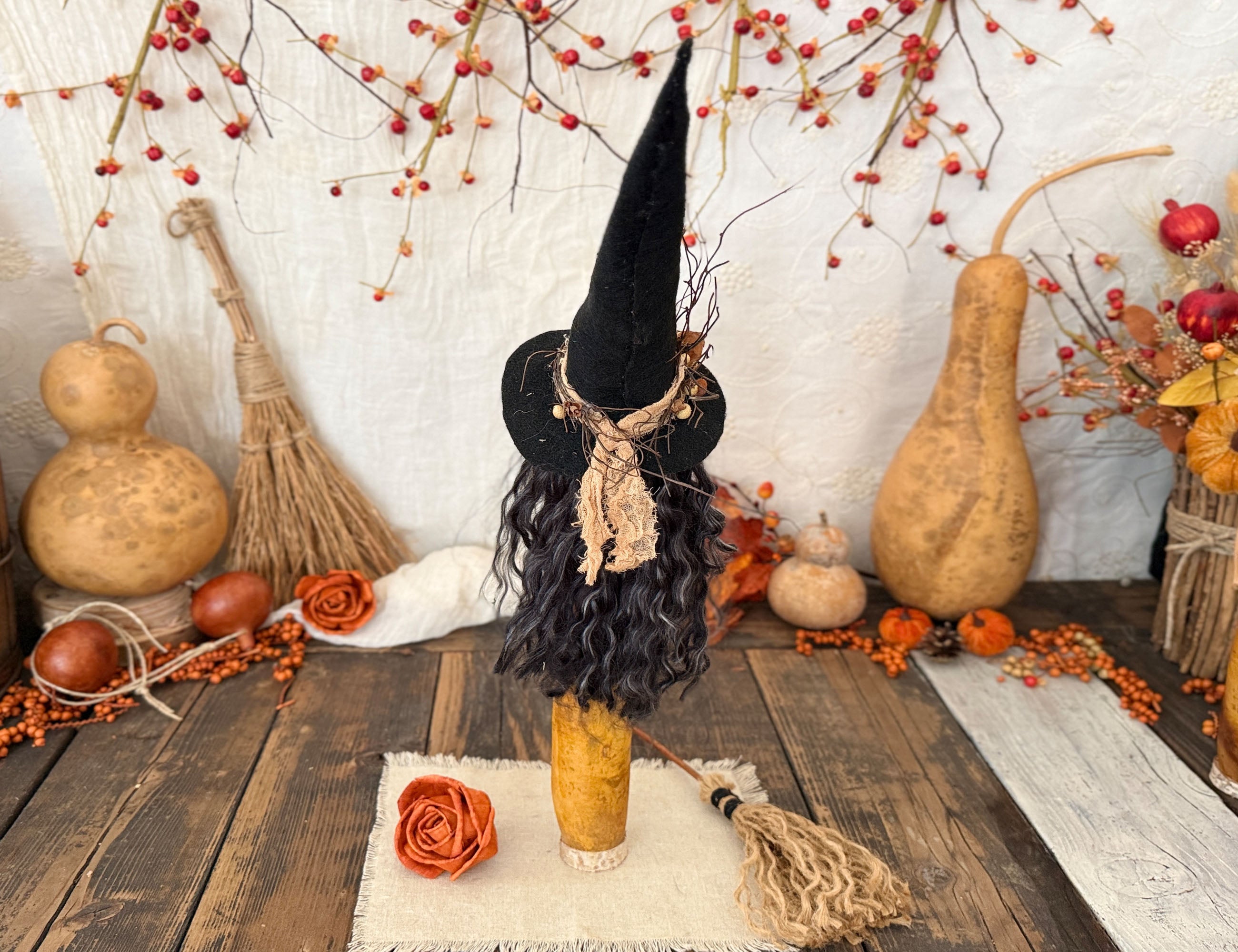 A Halloween witch decoration with a tall black hat, black curly hair, and beige scarf, displayed on a wooden surface with autumn props including gourds, pumpkins, a broom, and orange berries.