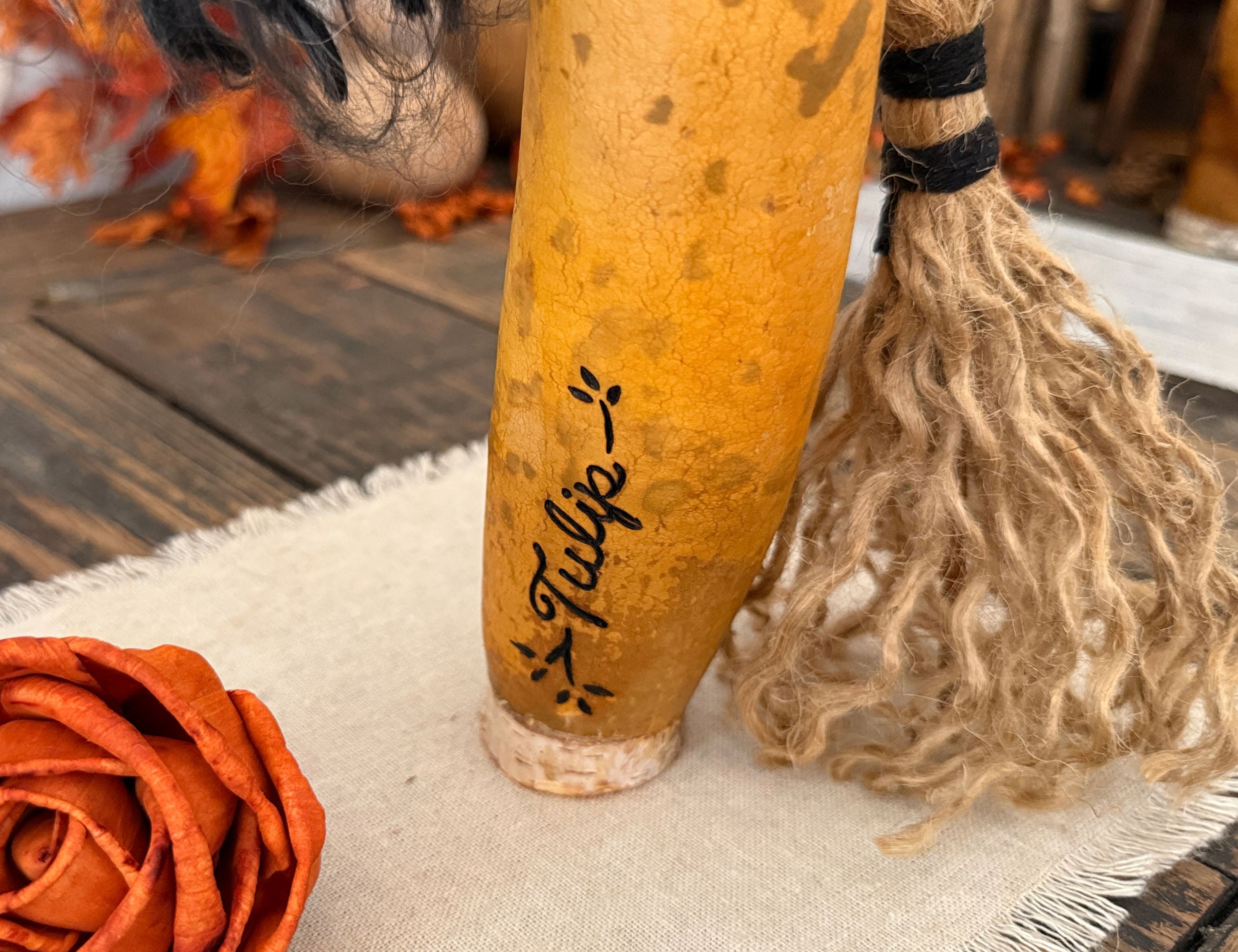 A close-up of a decorative gourd with handwritten black text reading 'Tulip' and small leaf accents, standing on a beige fabric surface next to a brown broom with tan bristles.