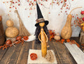 A decorative autumn witch figurine made from a gourd, wearing a black pointed hat and holding a broom, displayed on a rustic wooden surface with gourd props, orange berries, and a white textured backdrop.