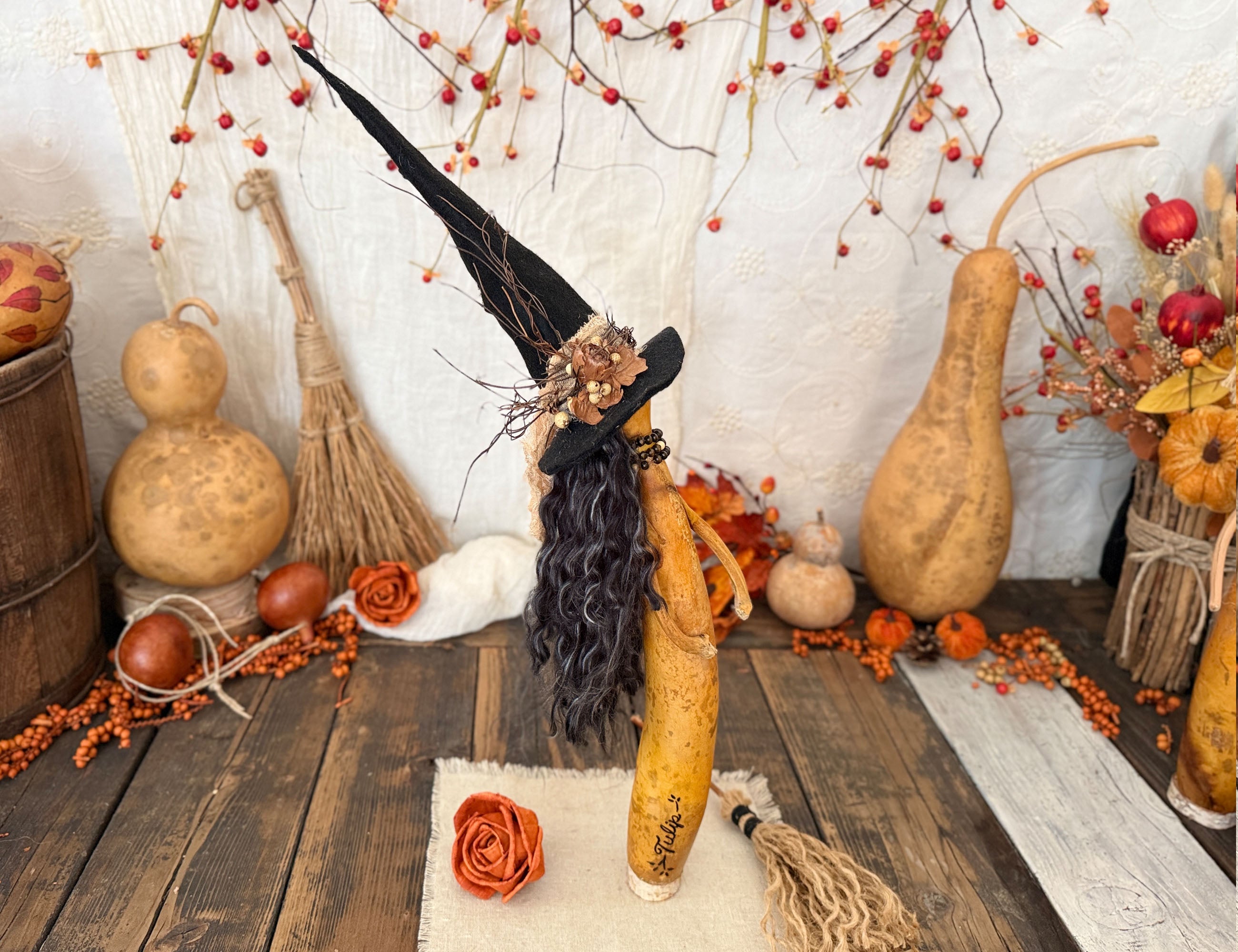 A decorative Halloween witch figure crafted from a gourd with long black yarn hair, a black pointed hat, and a small floral accent, displayed on a rustic wooden surface surrounded by autumn props including gourd vessels, berries, and brooms.