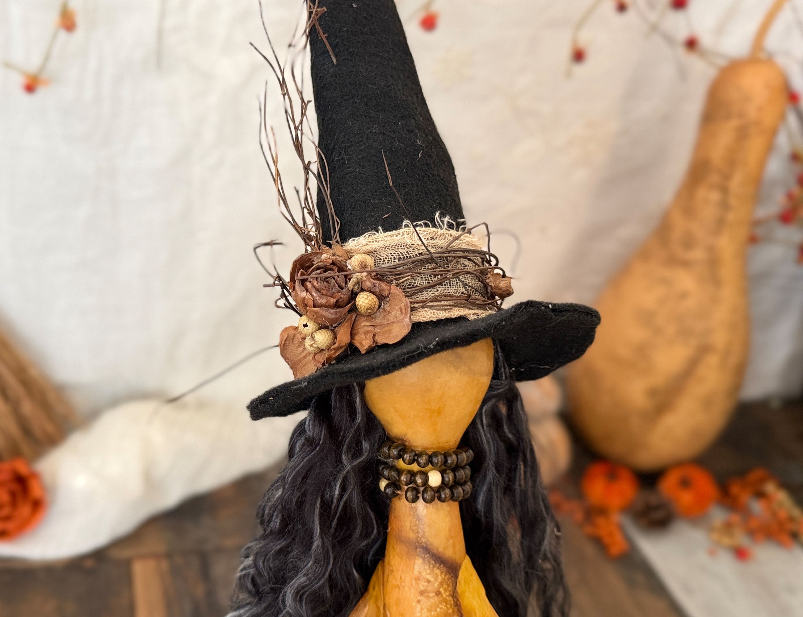 A decorative Halloween gourd doll with a black witch hat adorned with twigs and dried flowers, long black curly hair, and an orange-yellow carved body, shown in a styled fall setting with a gourd and pumpkins.