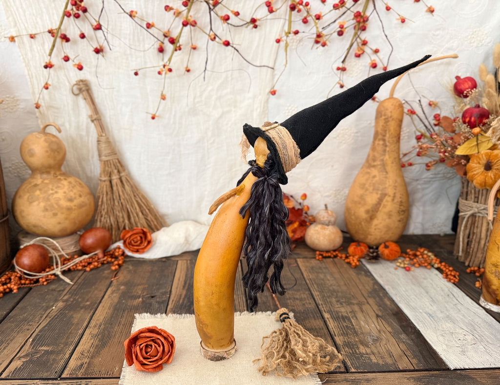 A decorative Halloween gourd sculpture shaped like a witch, made from a natural brown gourd with a black pointed hat, black hair and broom, displayed on a rustic wooden table with autumn decorations.