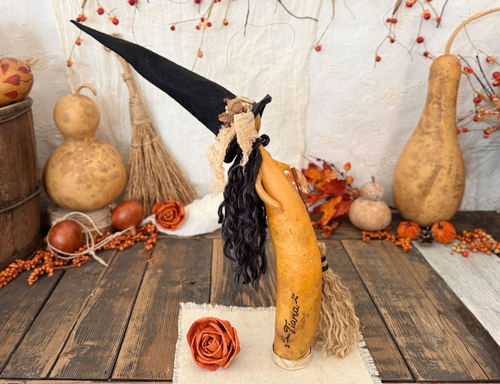A decorative carved gourd figure wearing a tall black witch hat and long black curly wig, posed on a rustic wooden surface with autumn props including gourd vases, orange berries, leaves, and small pumpkins.