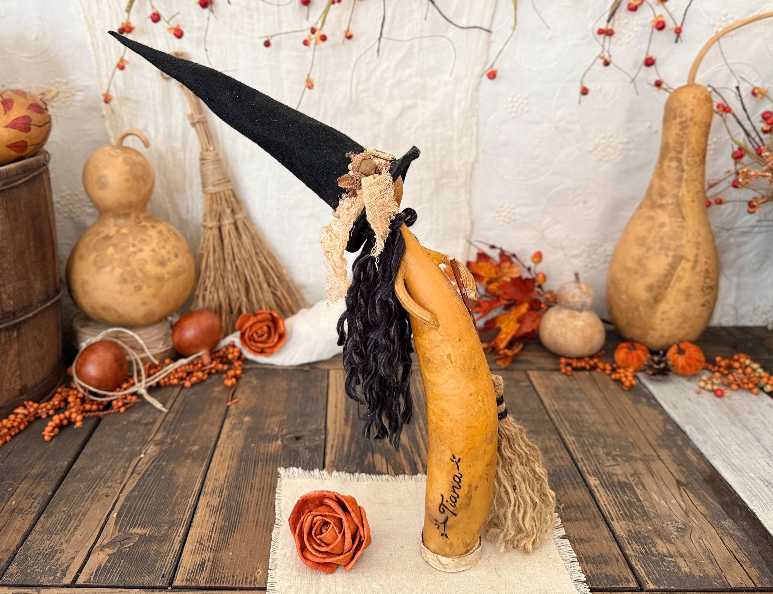 A decorative carved gourd figure wearing a tall black witch hat and long black curly wig, posed on a rustic wooden surface with autumn props including gourd vases, orange berries, leaves, and small pumpkins.