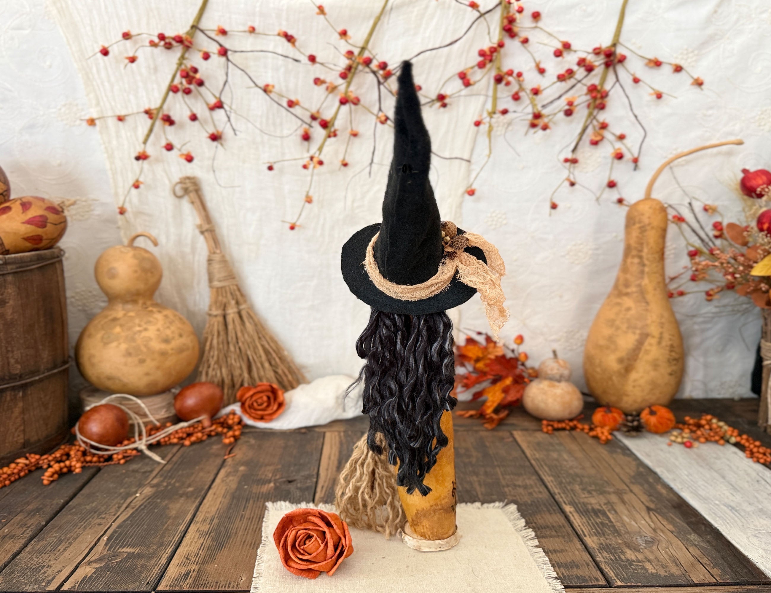 A Halloween witch decoration made from a hollow gourd with a tall black pointed hat, black curly hair, and brown broom, displayed on a rustic wooden table surrounded by autumn decorations.